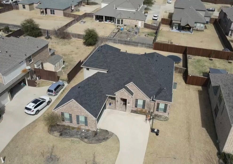 Aerial view of a suburban house with a dark roof, driveway, and parked white SUV in a neighborhood.