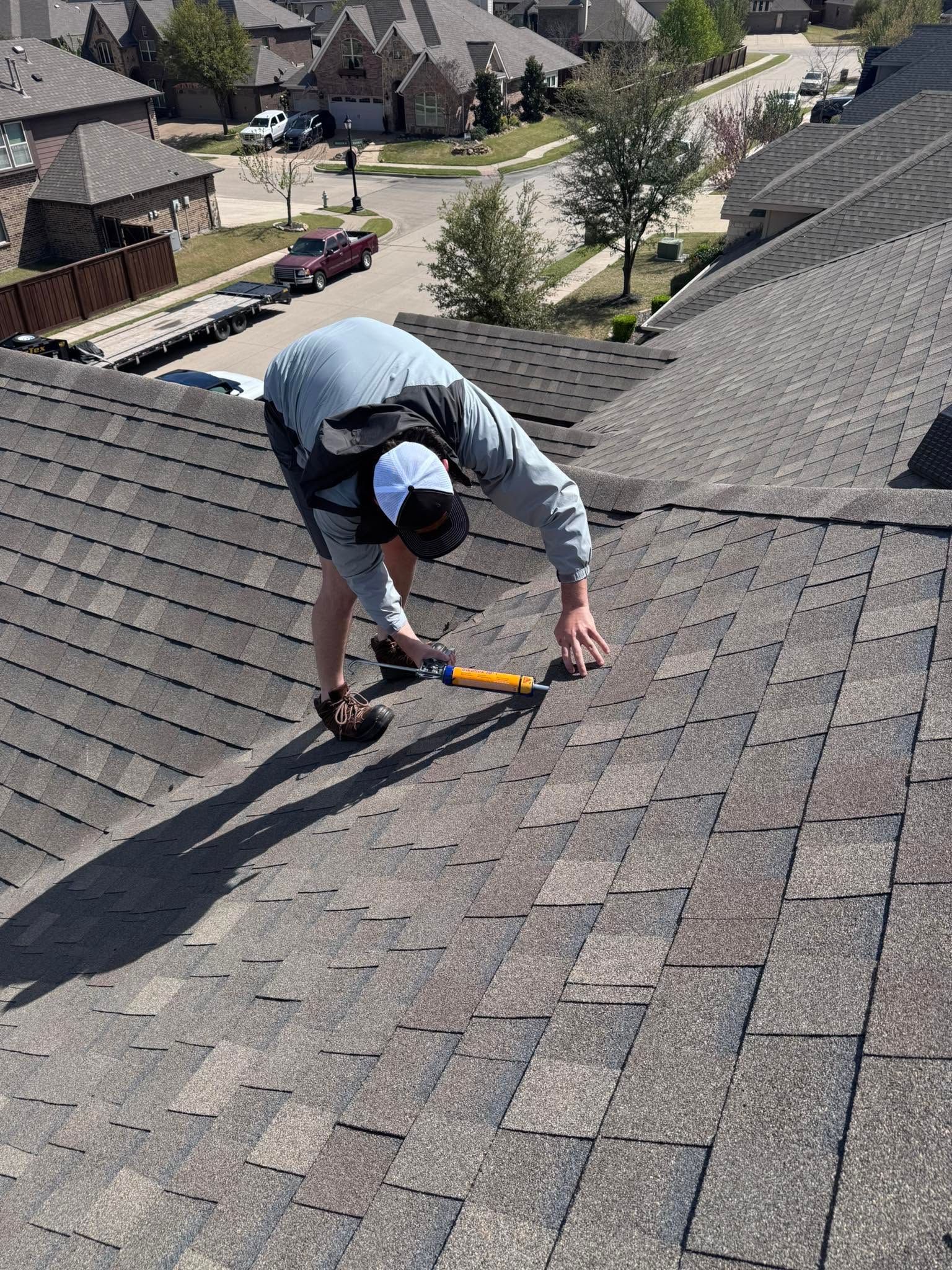Worker kneeling on a shingled roof, using a tool to inspect or repair shingles.