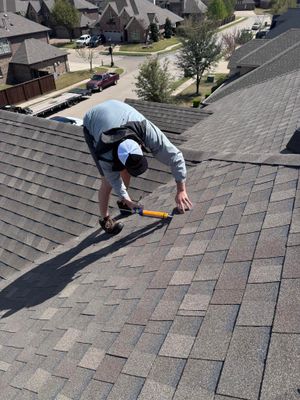 Worker kneeling on a shingled roof, using a tool to inspect or repair shingles.