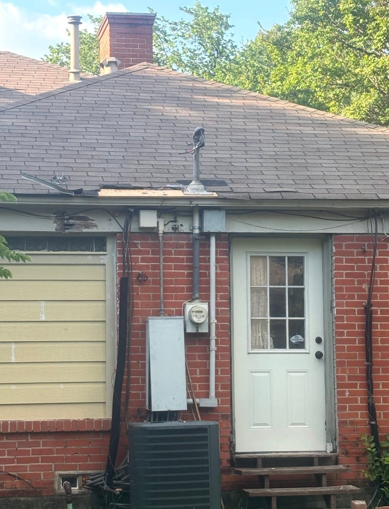 Brick house exterior with white door, garage door, utility boxes, and a shingled roof.