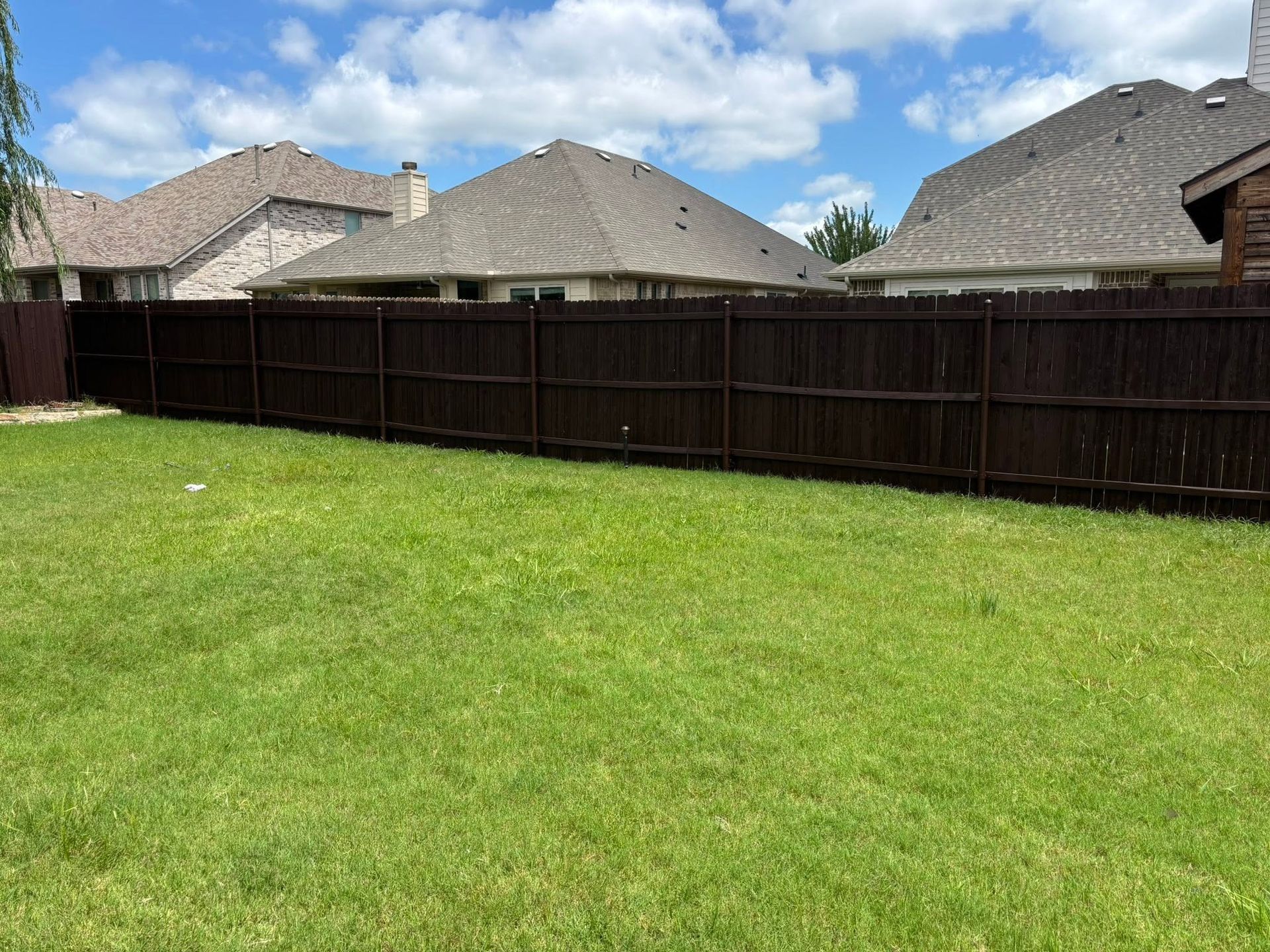 Grassy backyard with a dark wooden fence and suburban houses under a blue sky with clouds