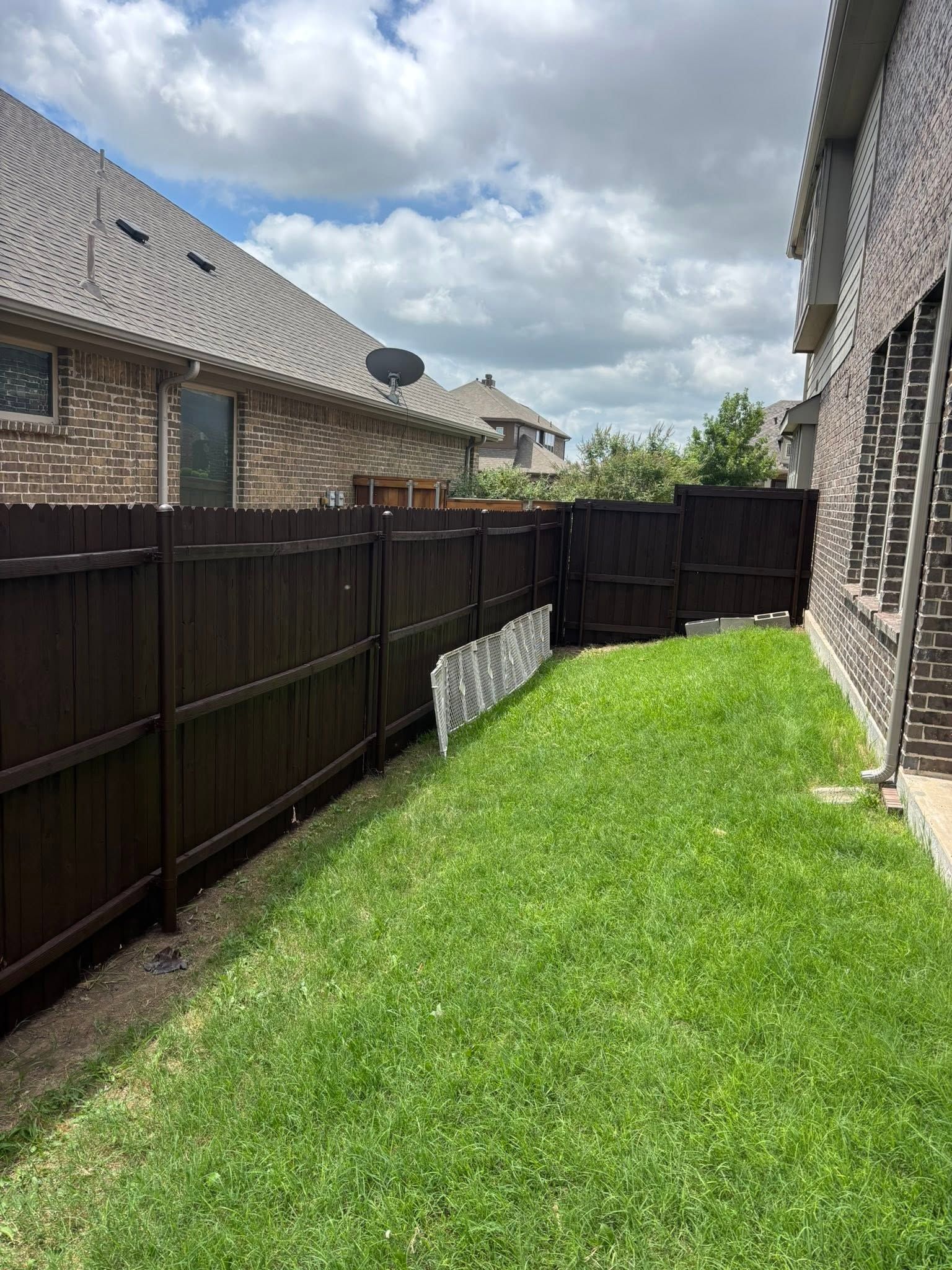 Narrow grassy backyard beside brick houses with a dark wooden fence under a cloudy sky