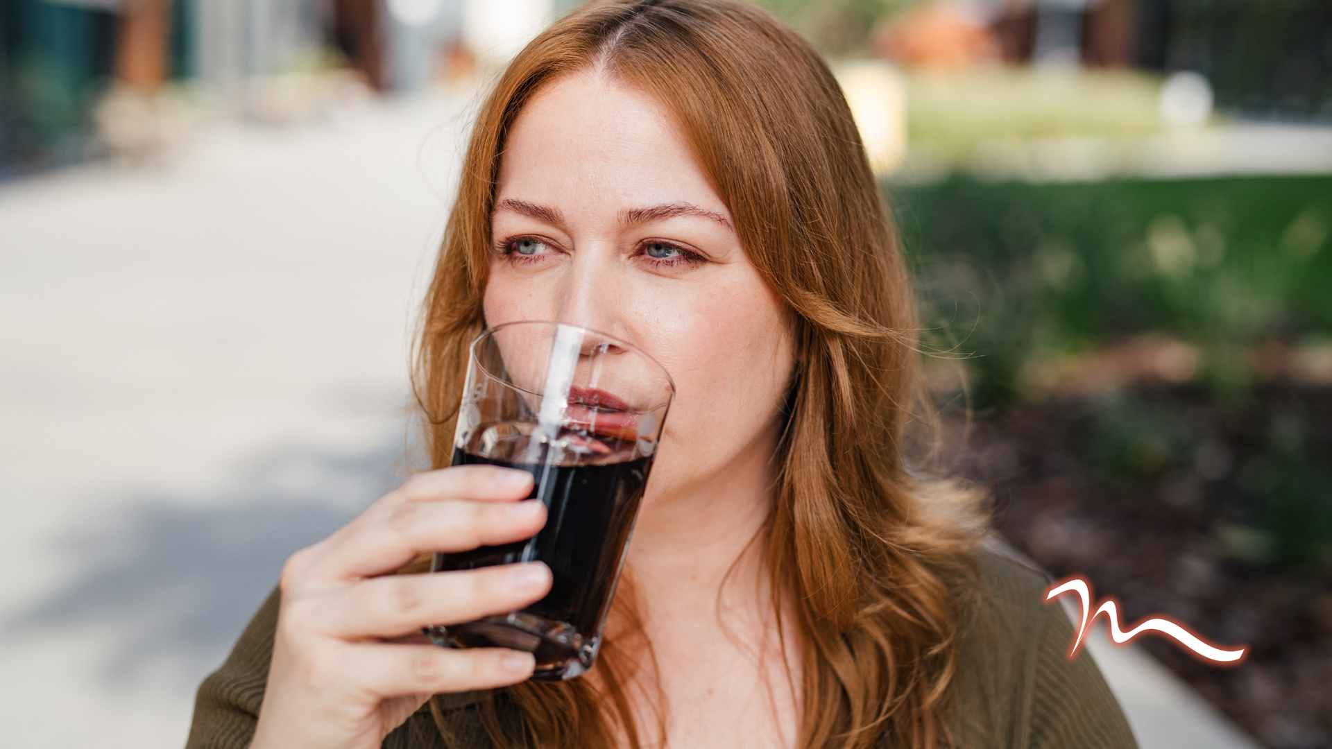 Woman outdoors drinking from a glass of dark beverage while smiling slightly.