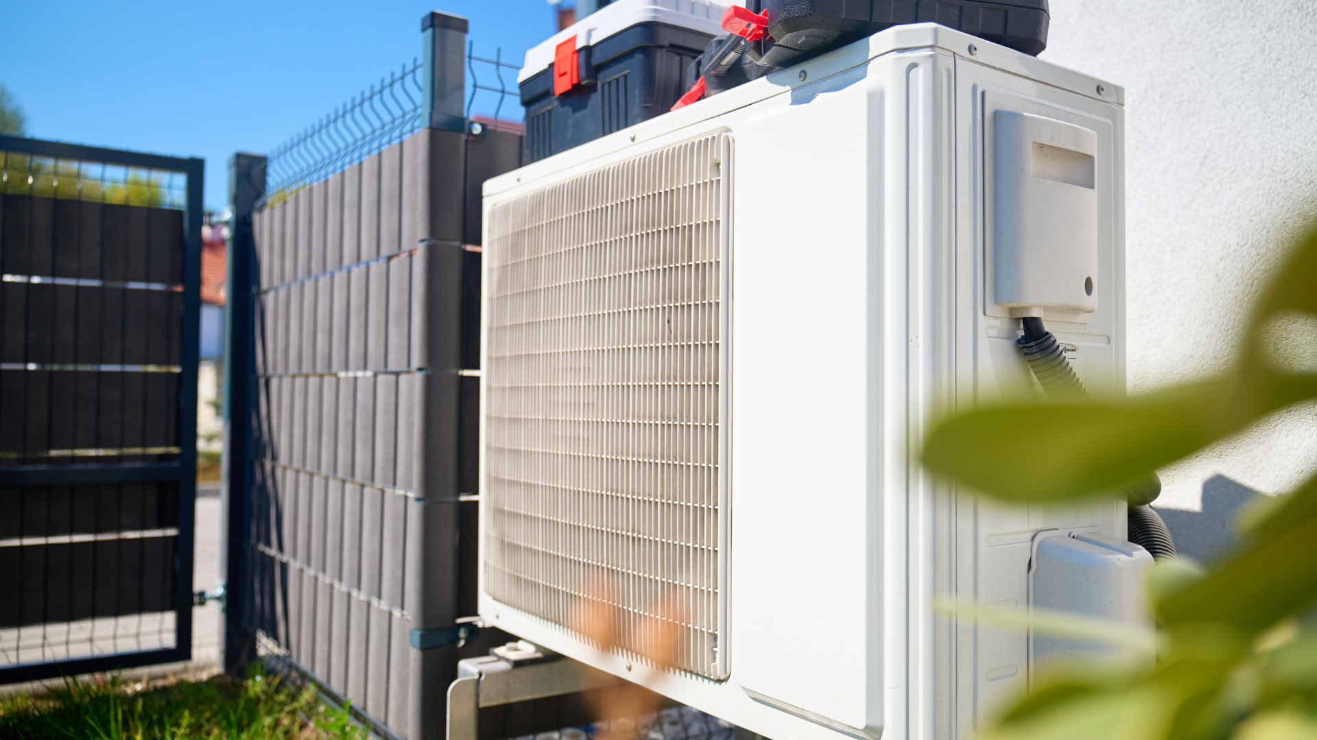 Outdoor HVAC unit beside a fence, with a white vented panel and leafy foreground.