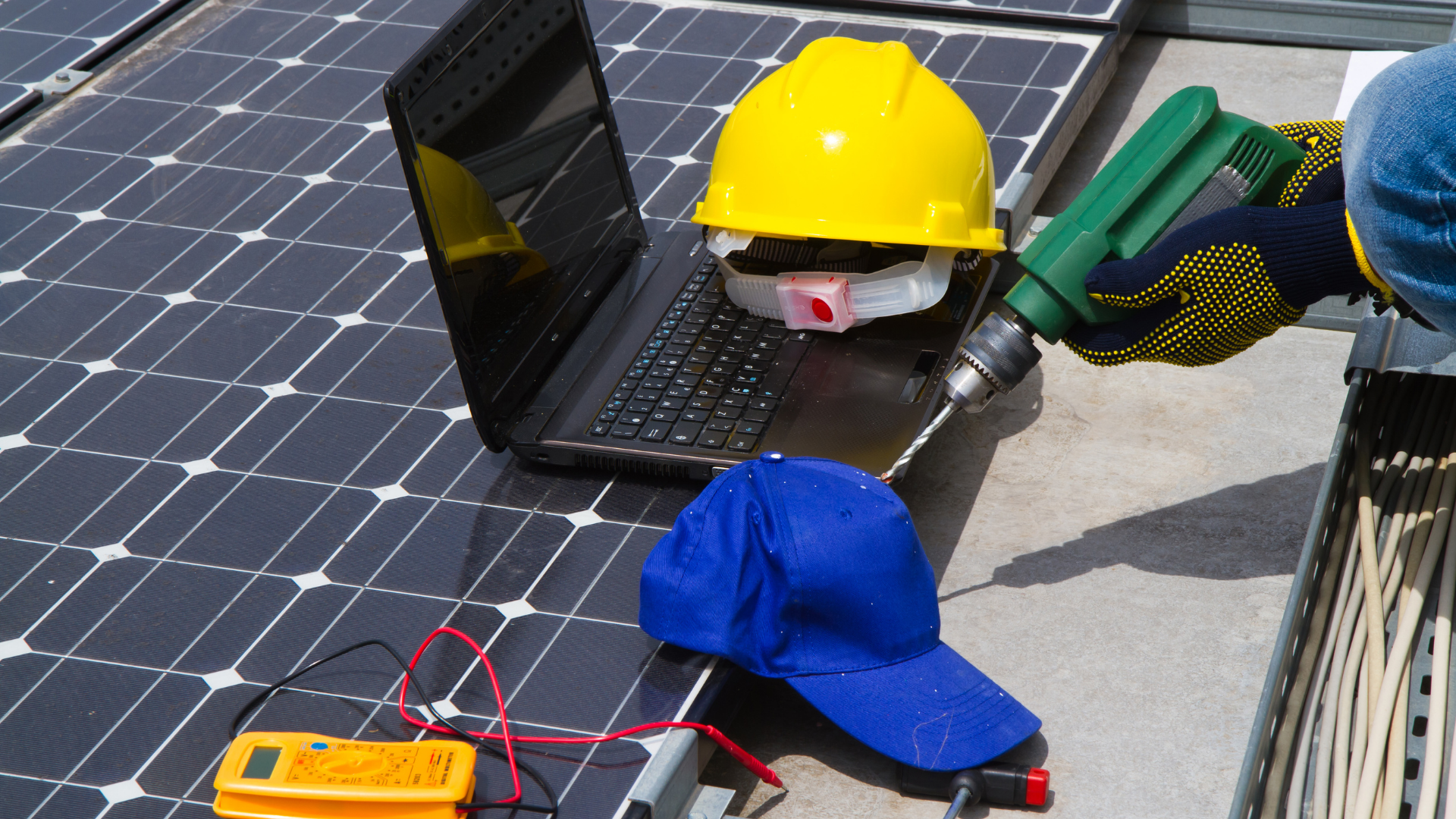 Solar panel maintenance tools beside a hard hat, laptop, gloves, and meter on a rooftop.