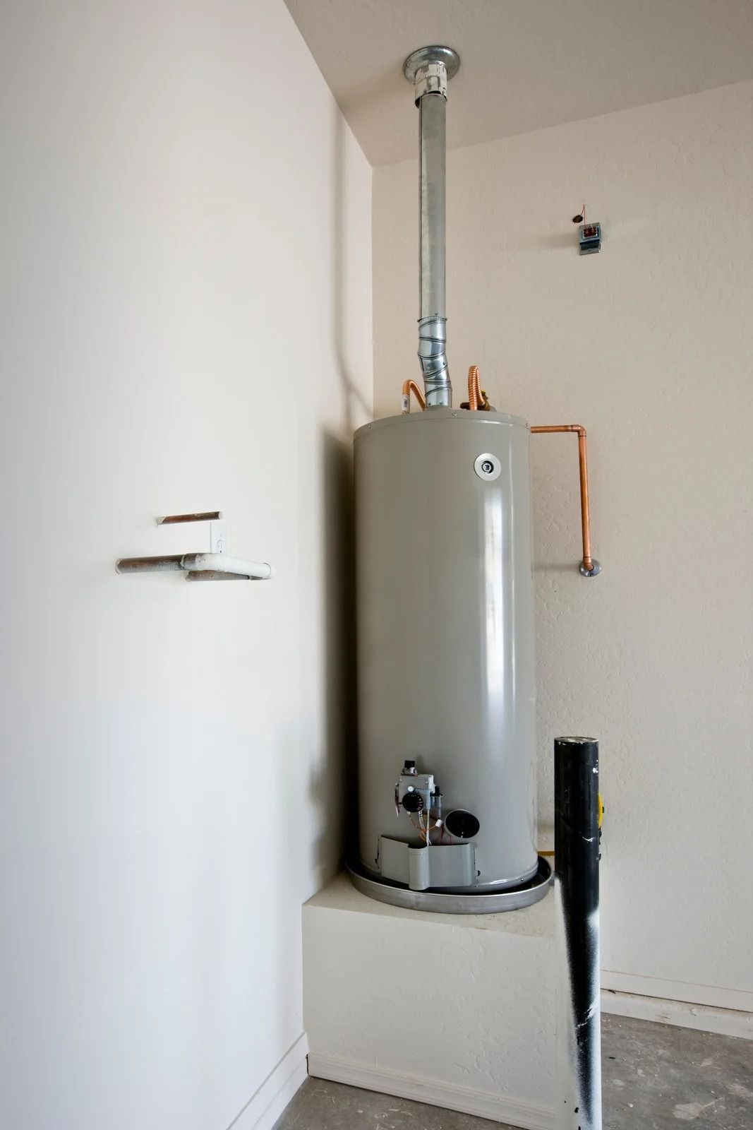 Tall gray water heater in a small utility closet with exposed pipes and white walls
