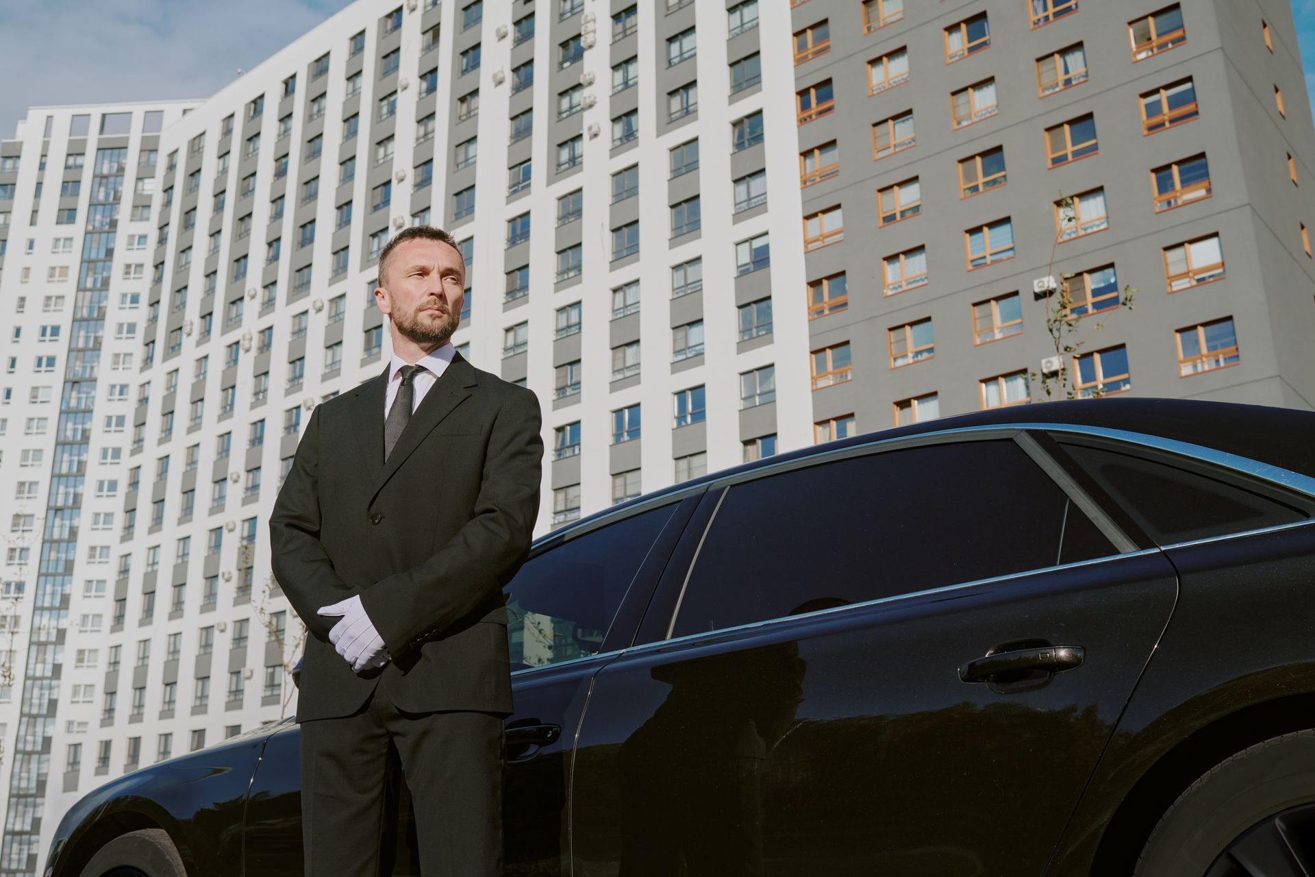 Man in a black suit stands beside a black car in front of a tall office building.