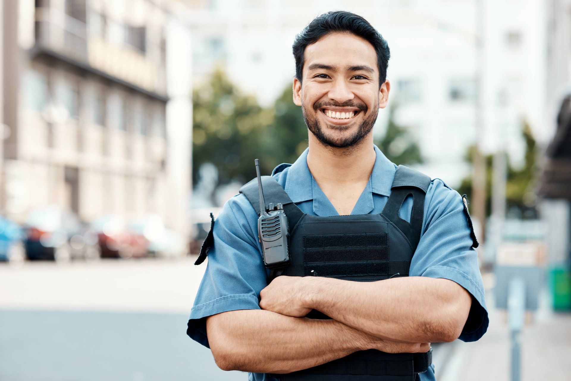 Smiling man with arms crossed, wearing a blue shirt and black vest, standing outdoors on a city street.