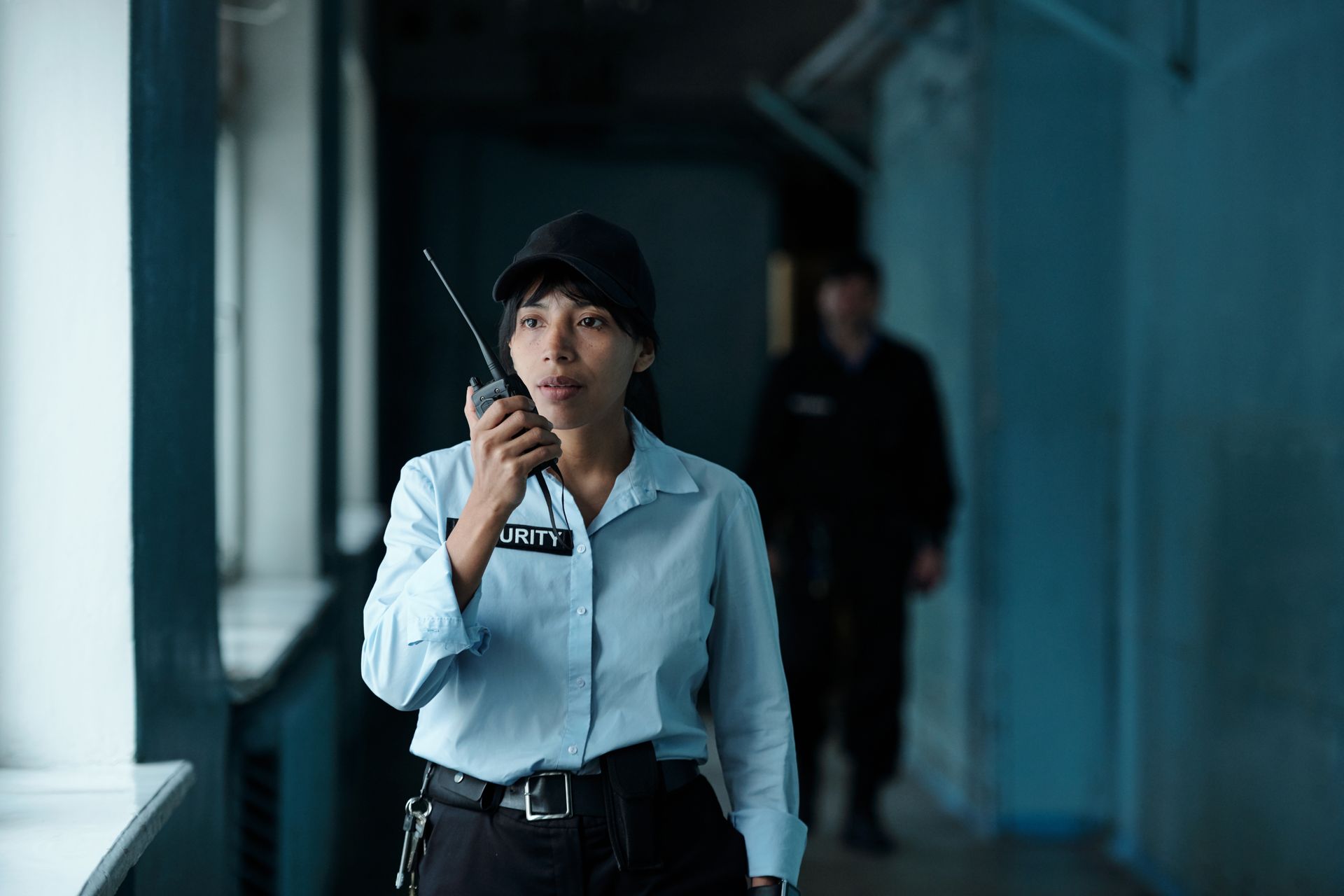 Woman in a light blue shirt speaking into a walkie-talkie in a dim hallway, with another figure behind her.