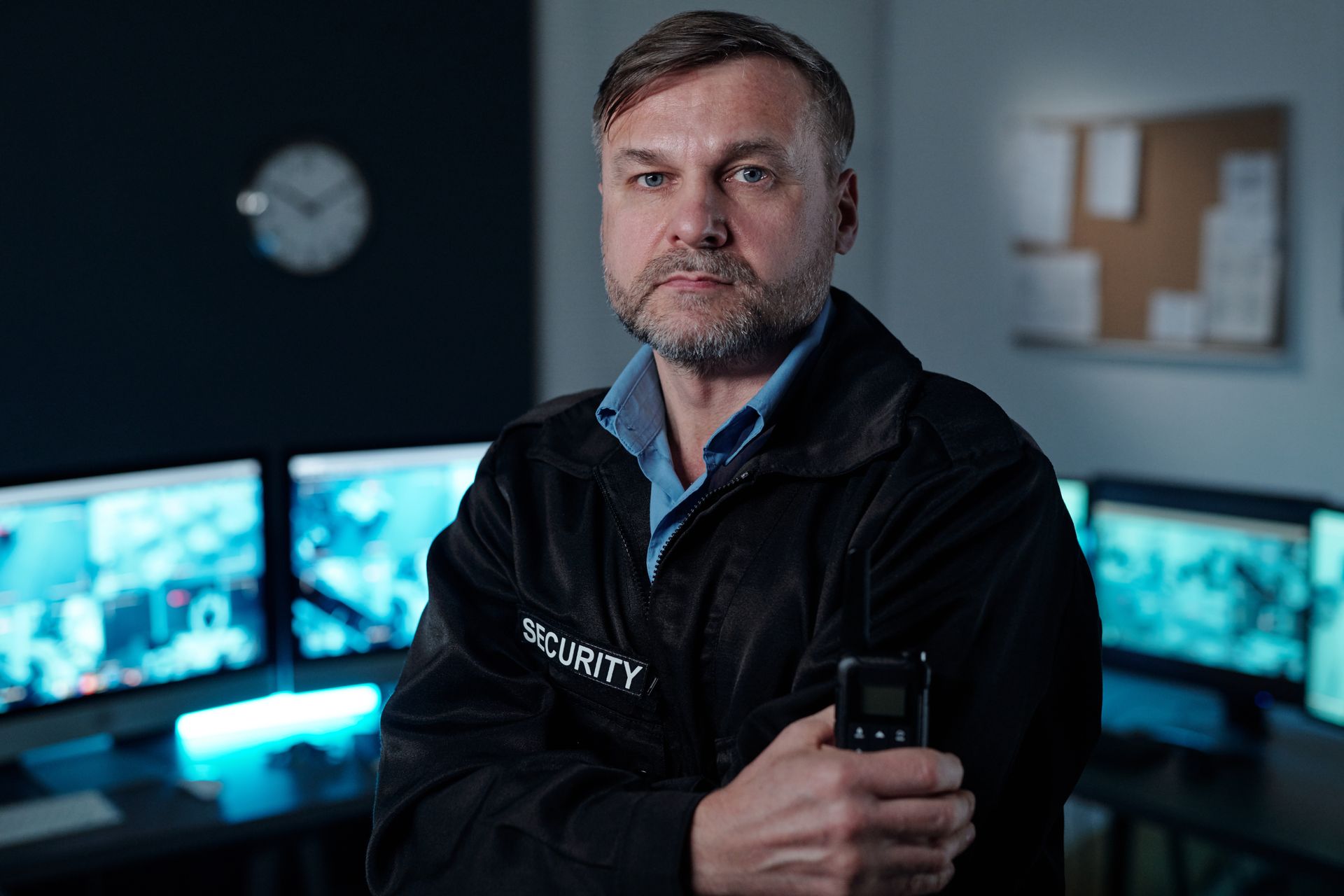 Security guard in a monitoring room, holding a phone with surveillance screens glowing behind him