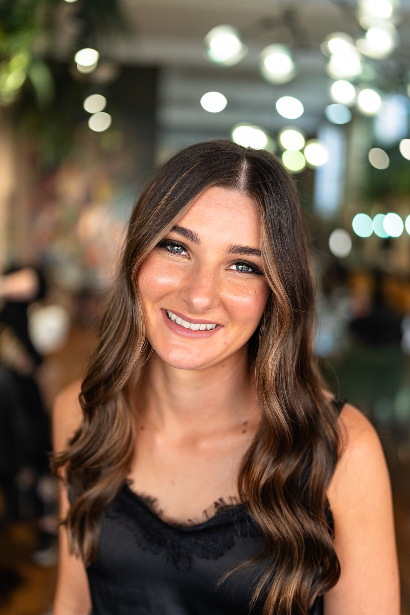 Woman with brown wavy hair smiling, wearing a black lace top, in a salon.