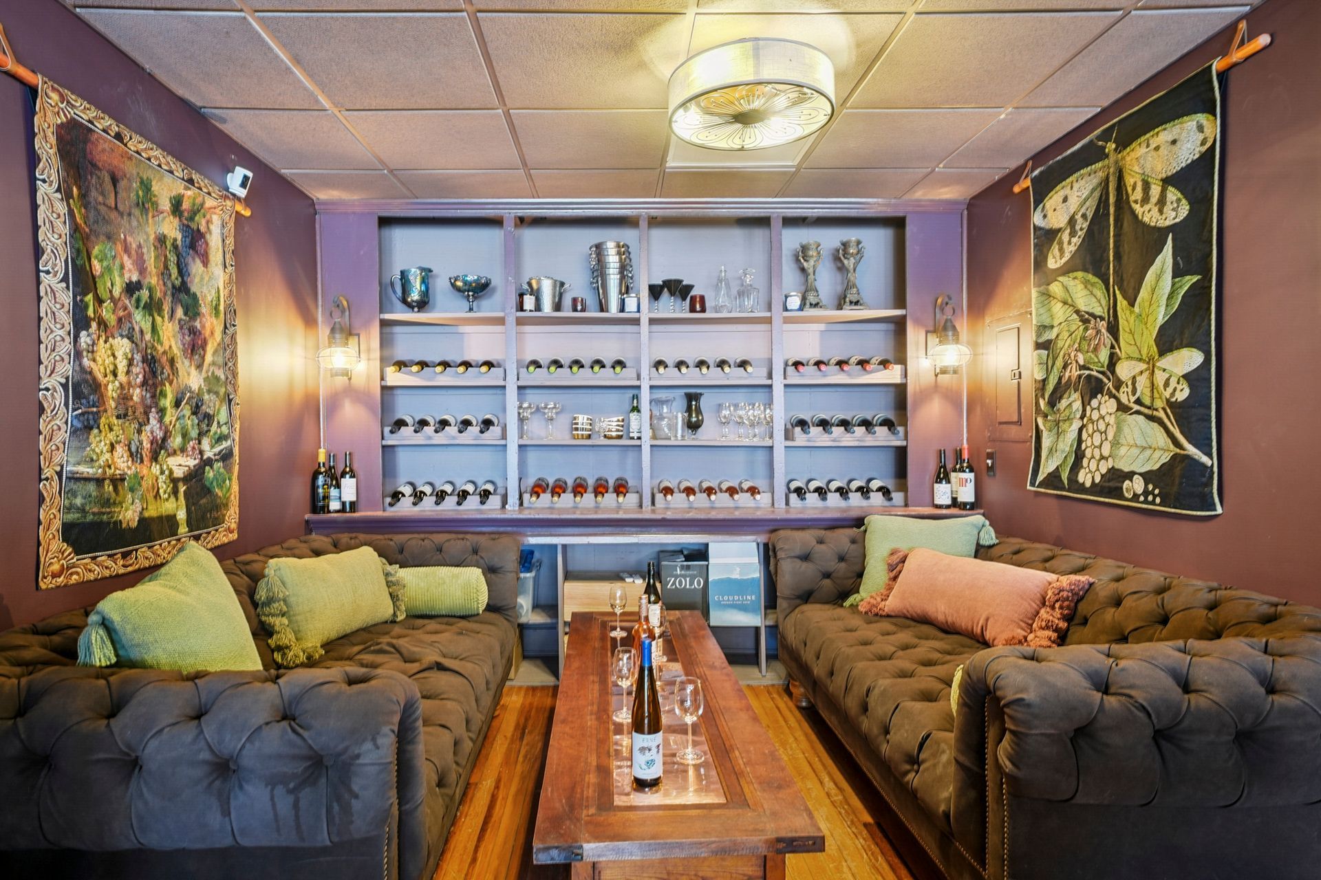 Cozy lounge with purple walls, tufted sofas, wall art, and shelves of bottles behind a central wooden table
