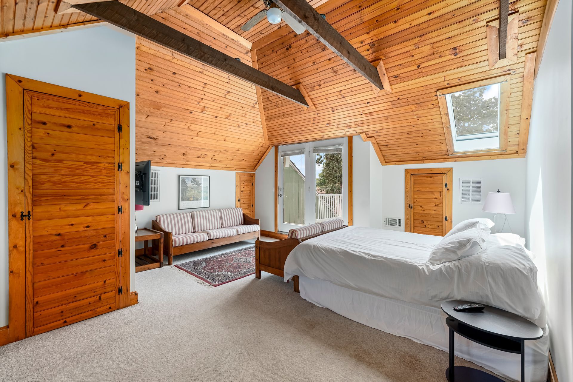 Cozy attic bedroom with wood-paneled ceiling, white bed, sofa, and large windows