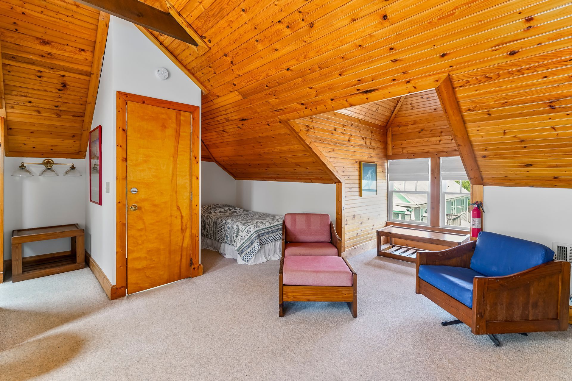 Cozy attic bedroom with wood ceiling, bed, pink chair, blue armchair, and large window.