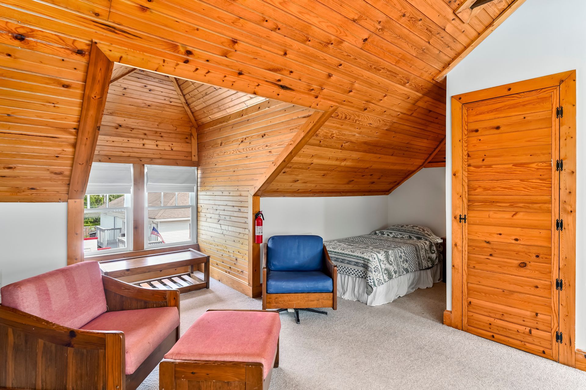 Cozy attic bedroom with wood-paneled vaulted ceiling, pink seating, blue chair, bed, and large windows