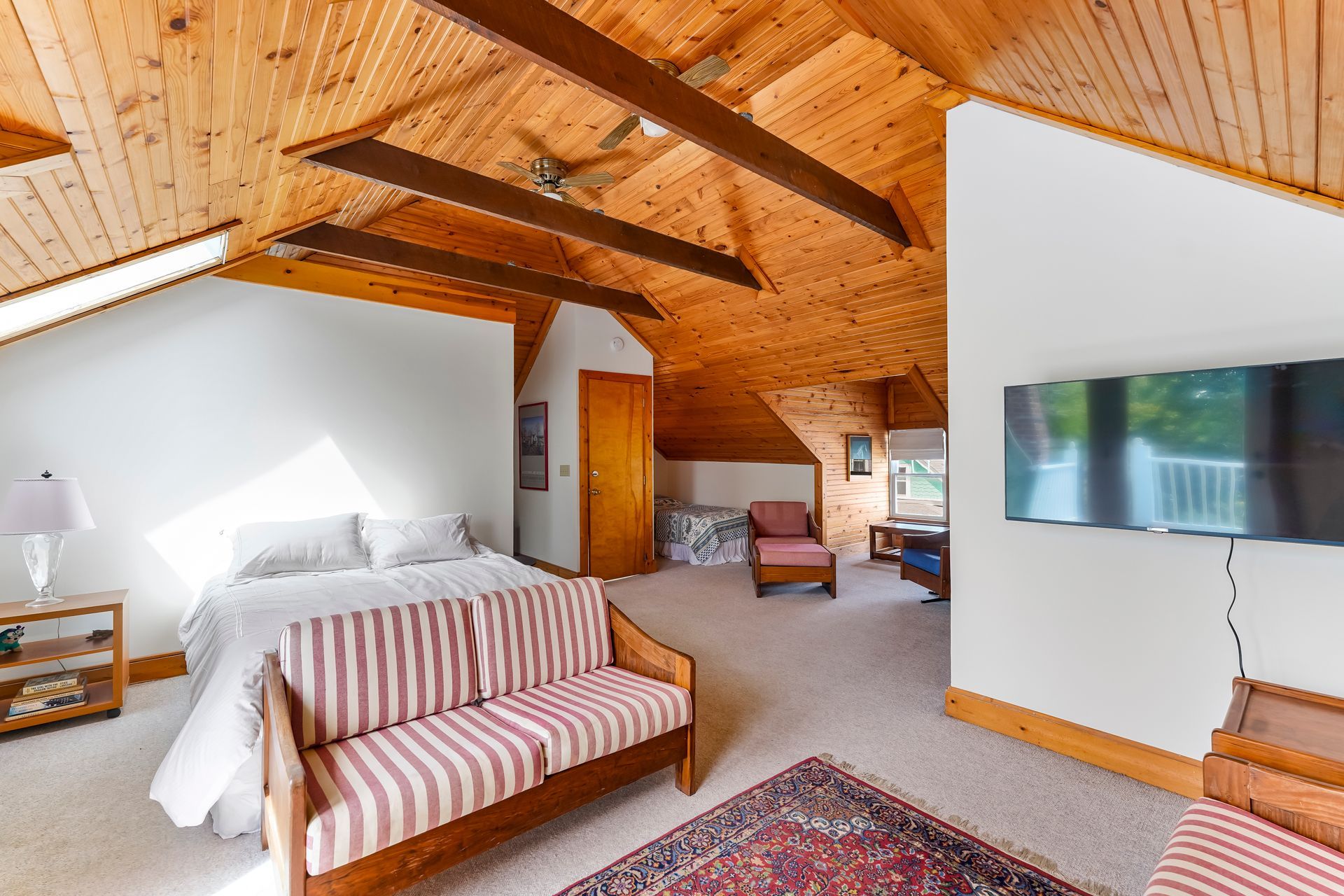 Spacious attic bedroom with wood-paneled ceiling, white bed, sofa, and wall-mounted TV