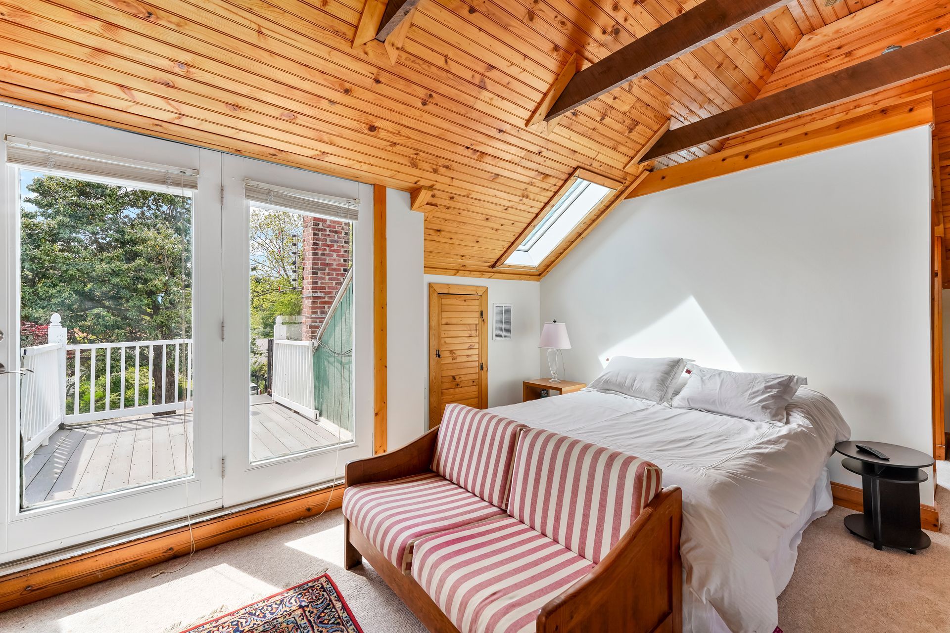 Cozy attic bedroom with wooden ceiling, white bed, red-striped sofa, and a balcony view