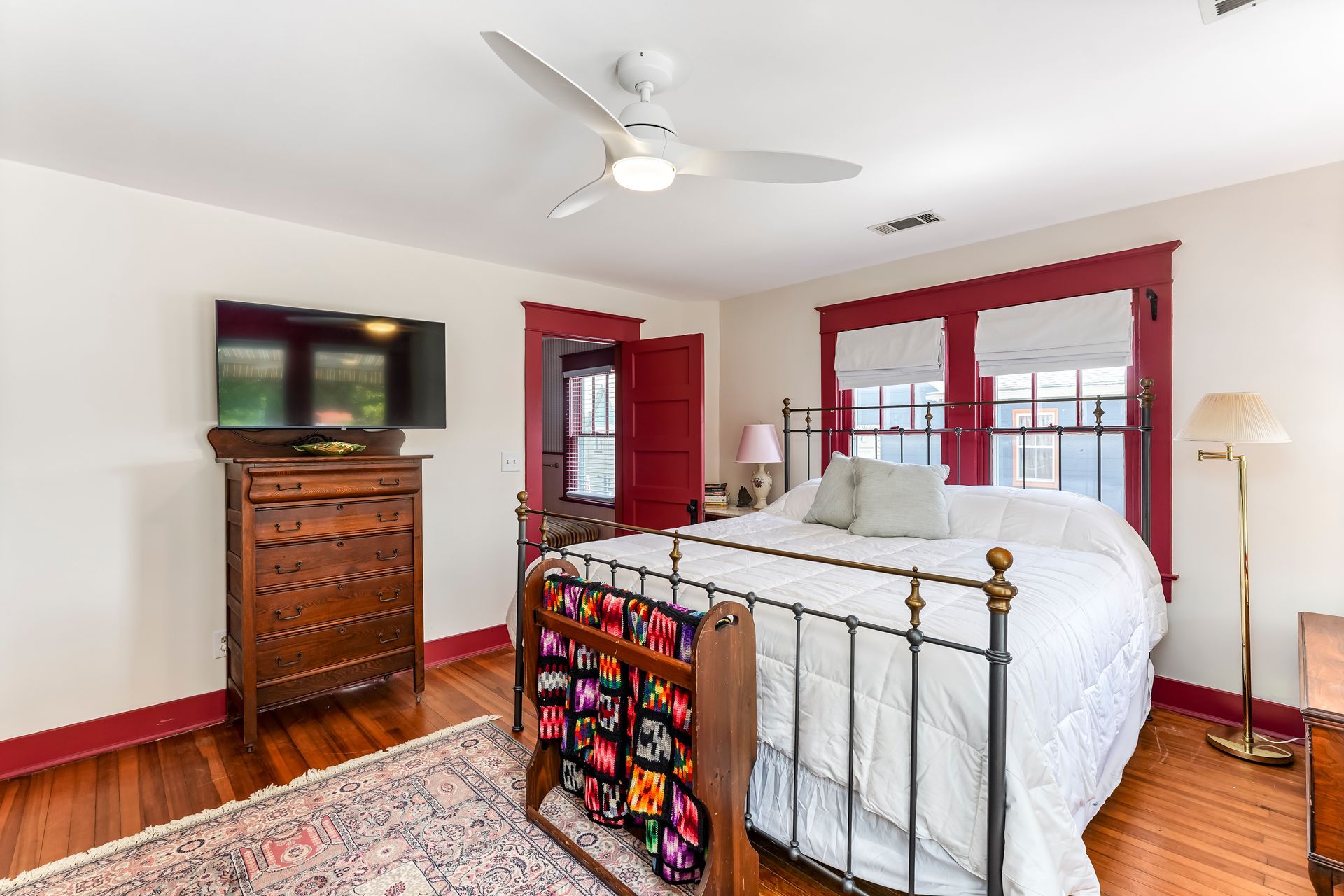 Bright bedroom with brass bed, red trim, wooden dresser, TV, and large windows