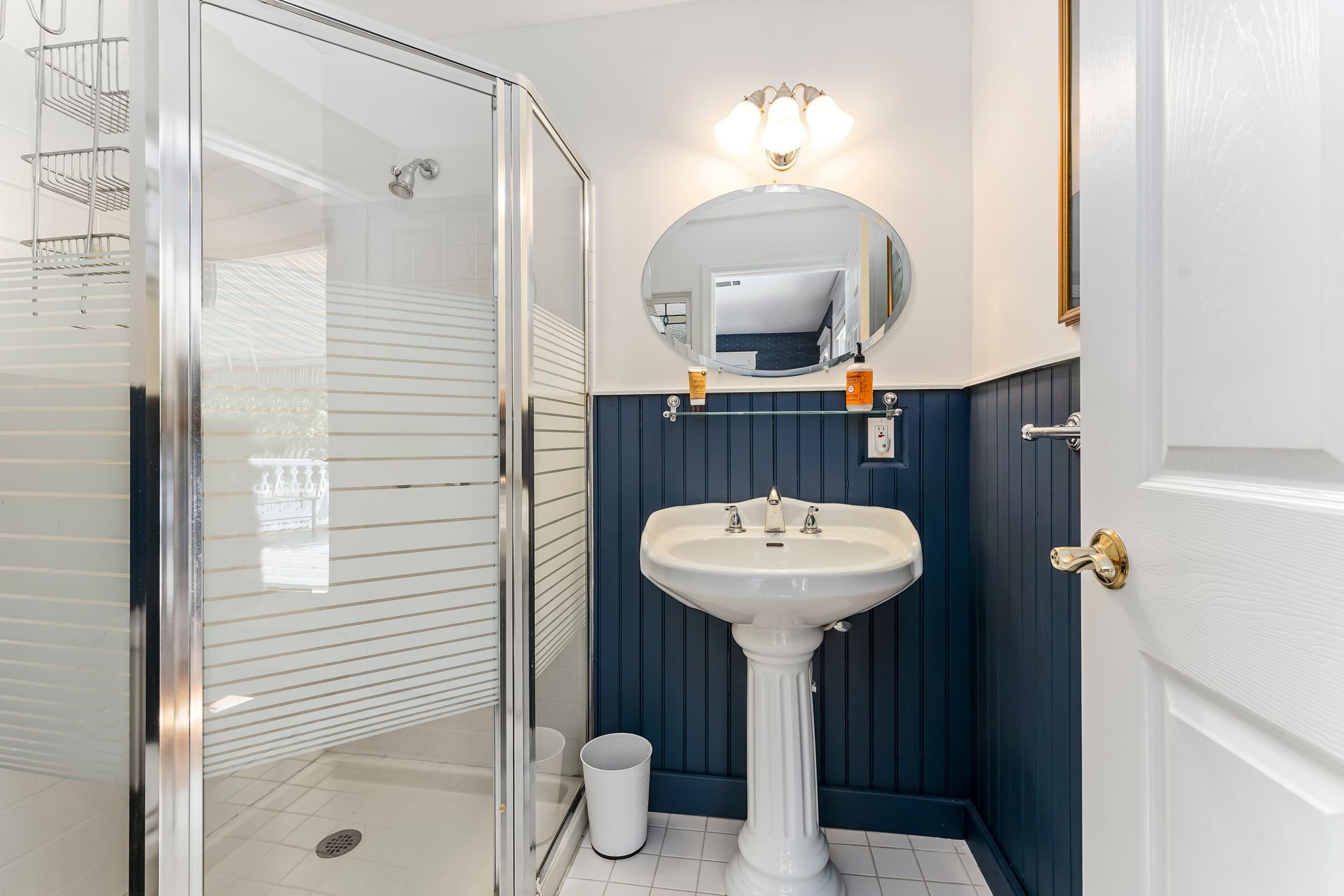 Small bathroom with pedestal sink, round mirror, glass shower, and navy wainscoting.