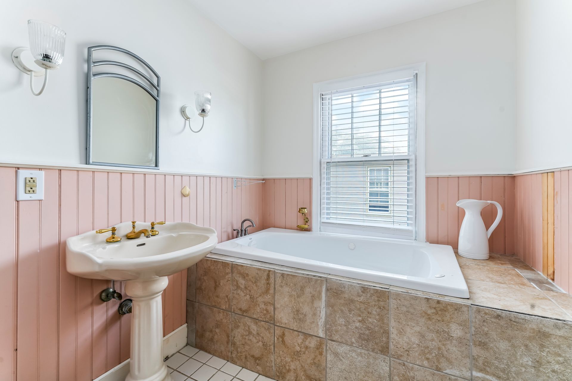 Bright bathroom with pink wainscoting, pedestal sink, and a tiled bathtub under a window.