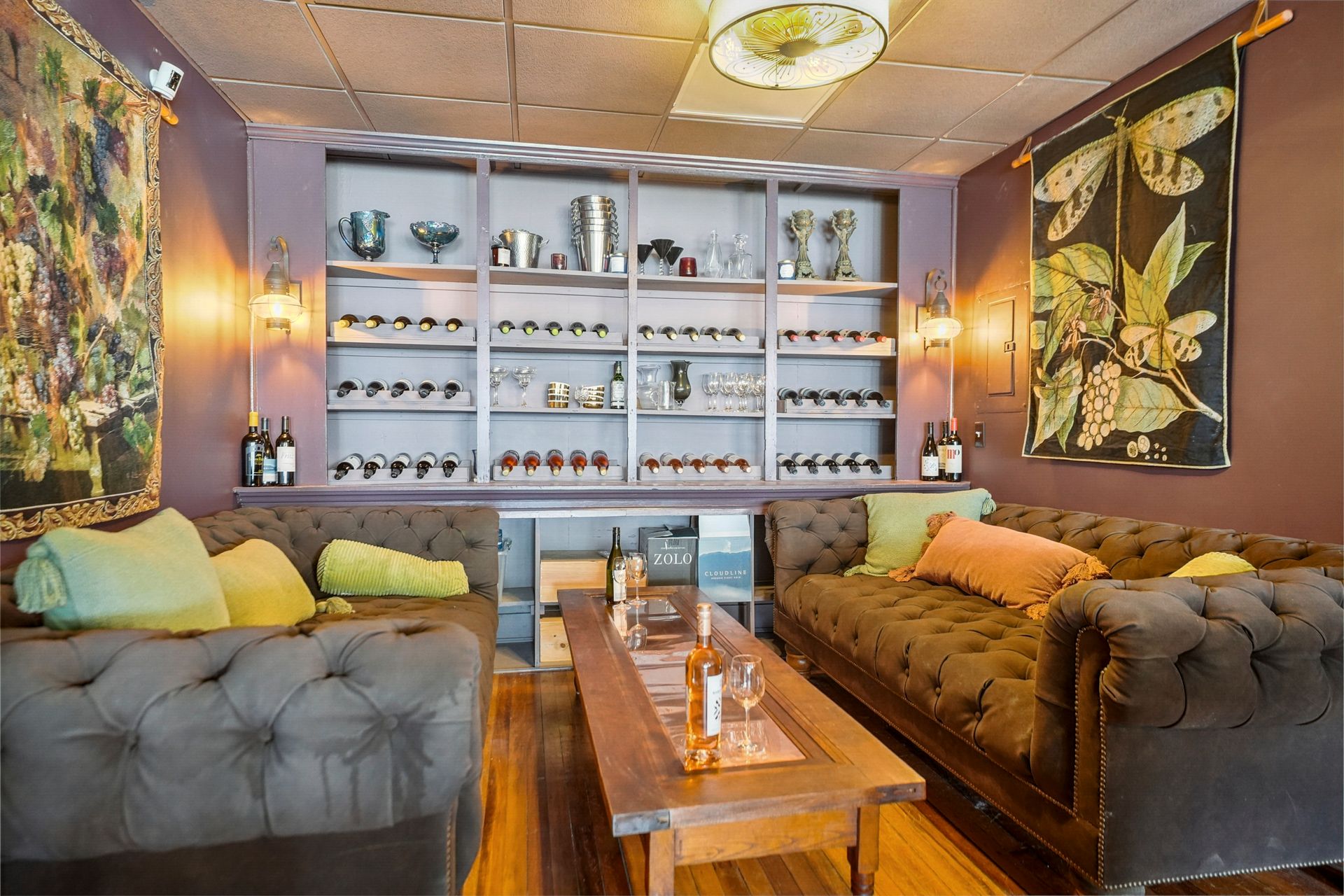 Cozy lounge with tufted sofas, wooden table, and wall shelves of bottles under warm lighting