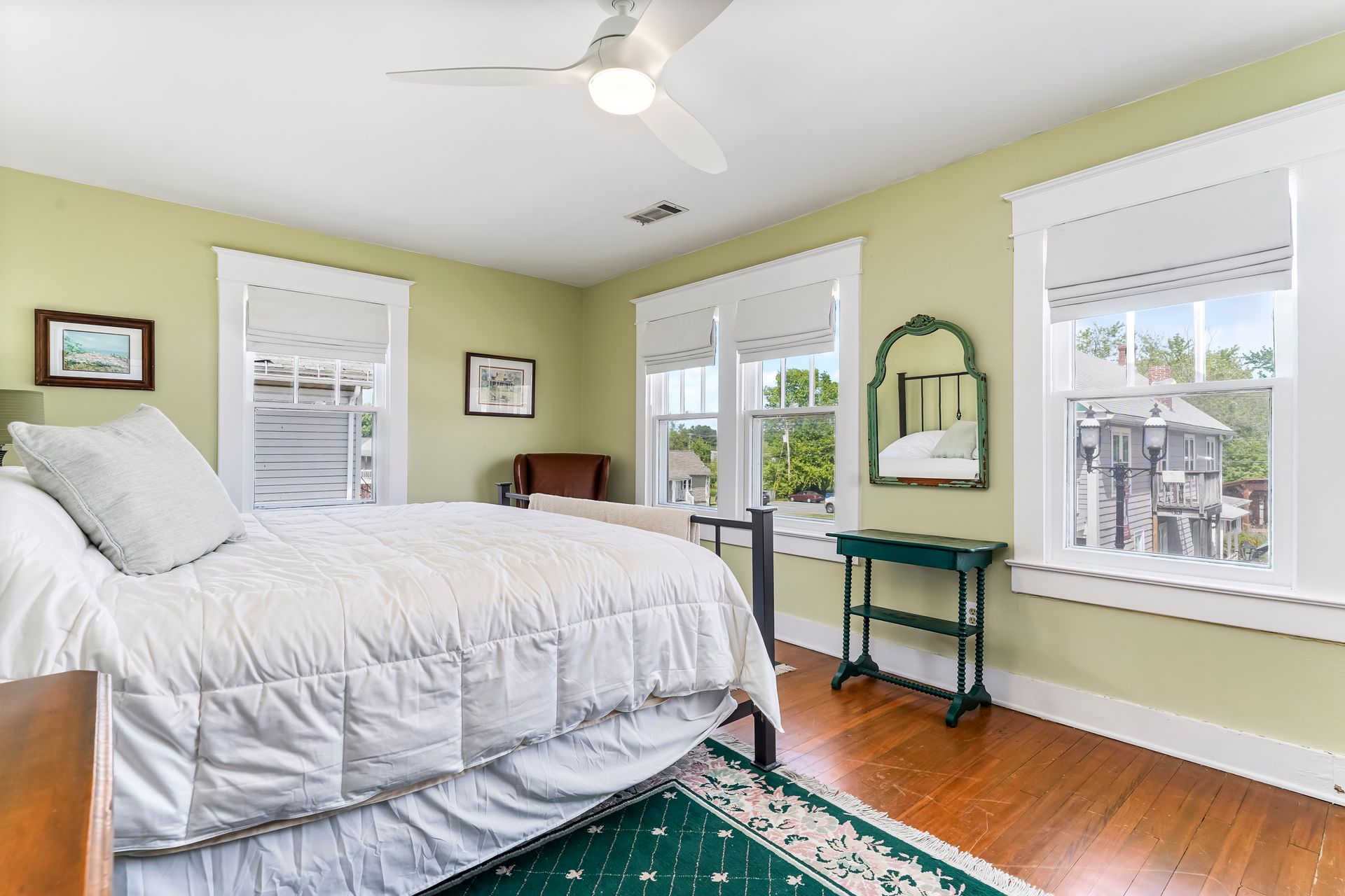 Bright bedroom with white bed, green walls, hardwood floor, and large windows with a desk by one window