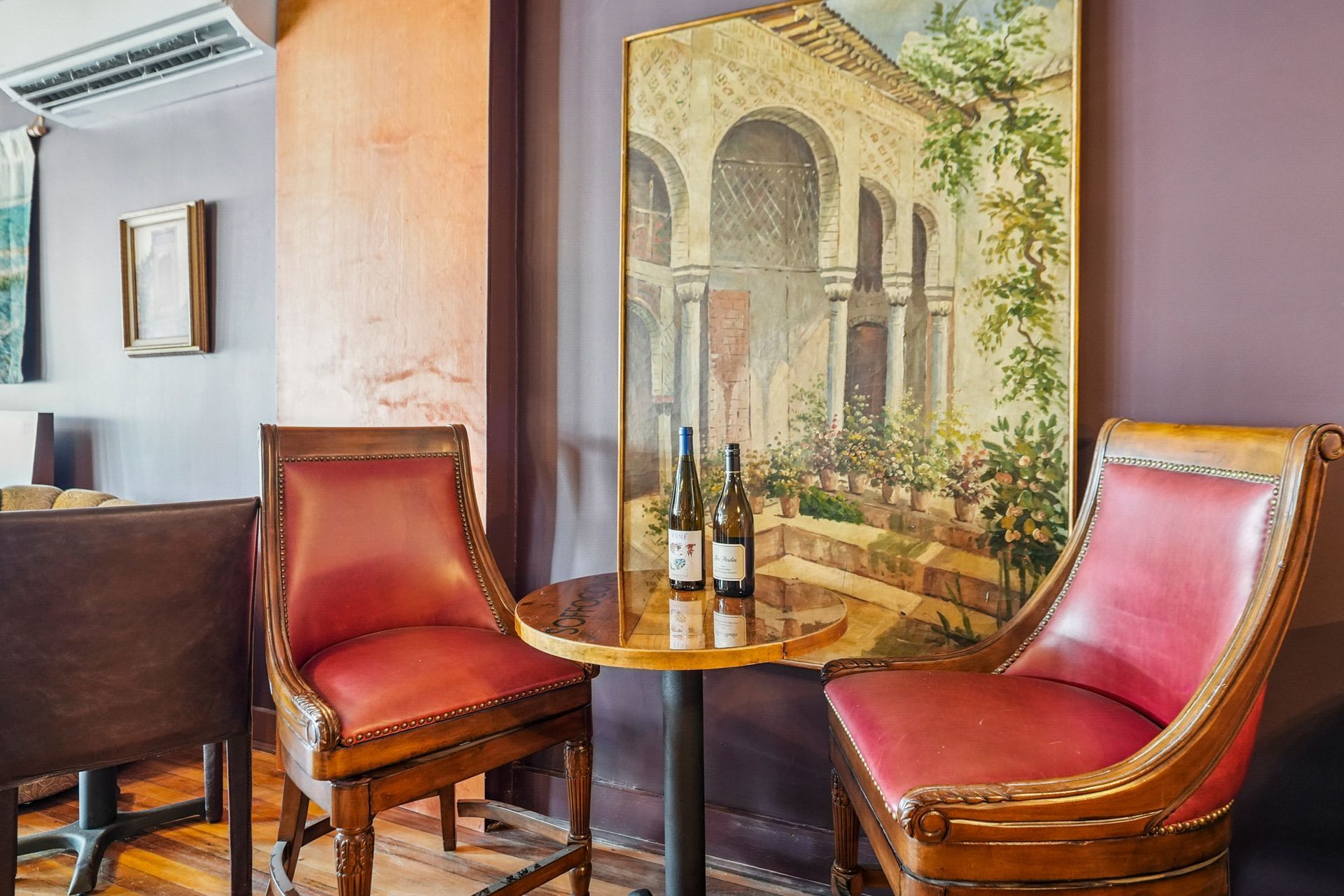 Elegant lounge with red leather chairs, round table, wine bottles, and a framed mural of an Italian courtyard