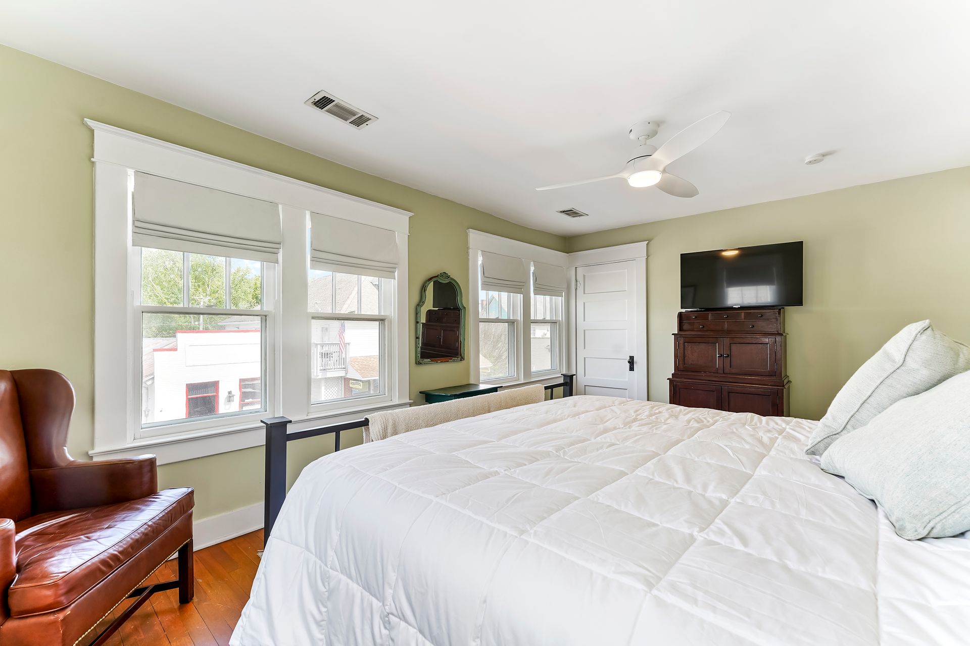 Bright bedroom with white bed, leather chair, windows, ceiling fan, and dresser with TV