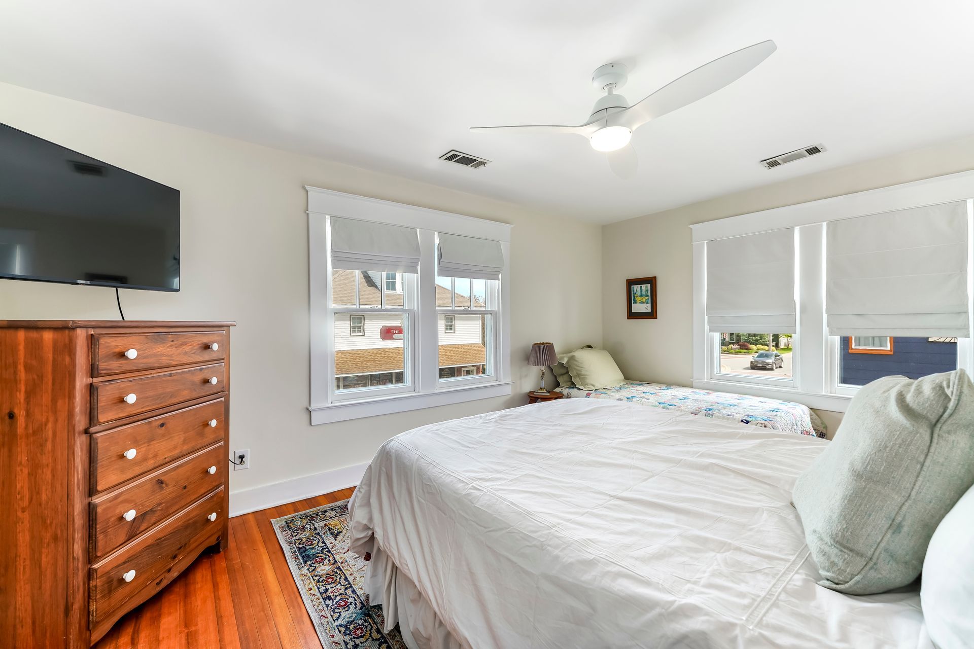 Bright bedroom with a white bed, wood dresser, wall TV, and two windows with blinds.
