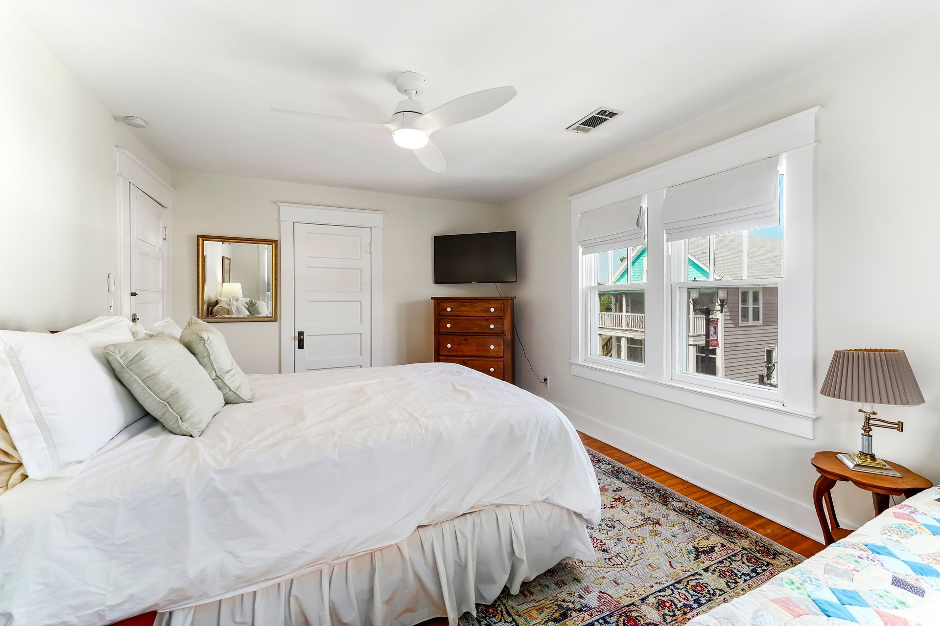 Bright bedroom with white walls, bed, dresser, TV, patterned rug, and large windows