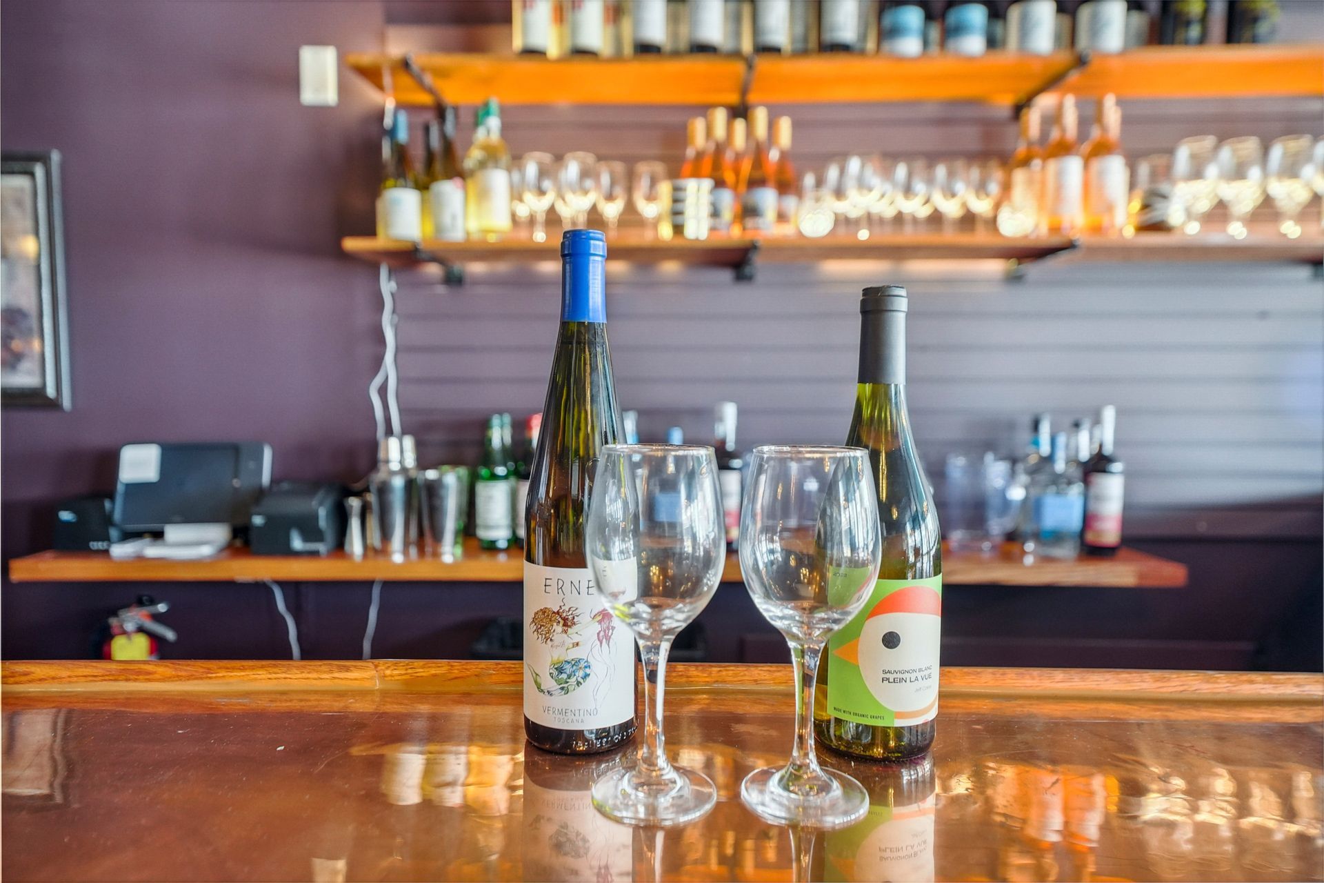 Two wine bottles and two empty glasses on a bar counter with shelves of liquor in the background