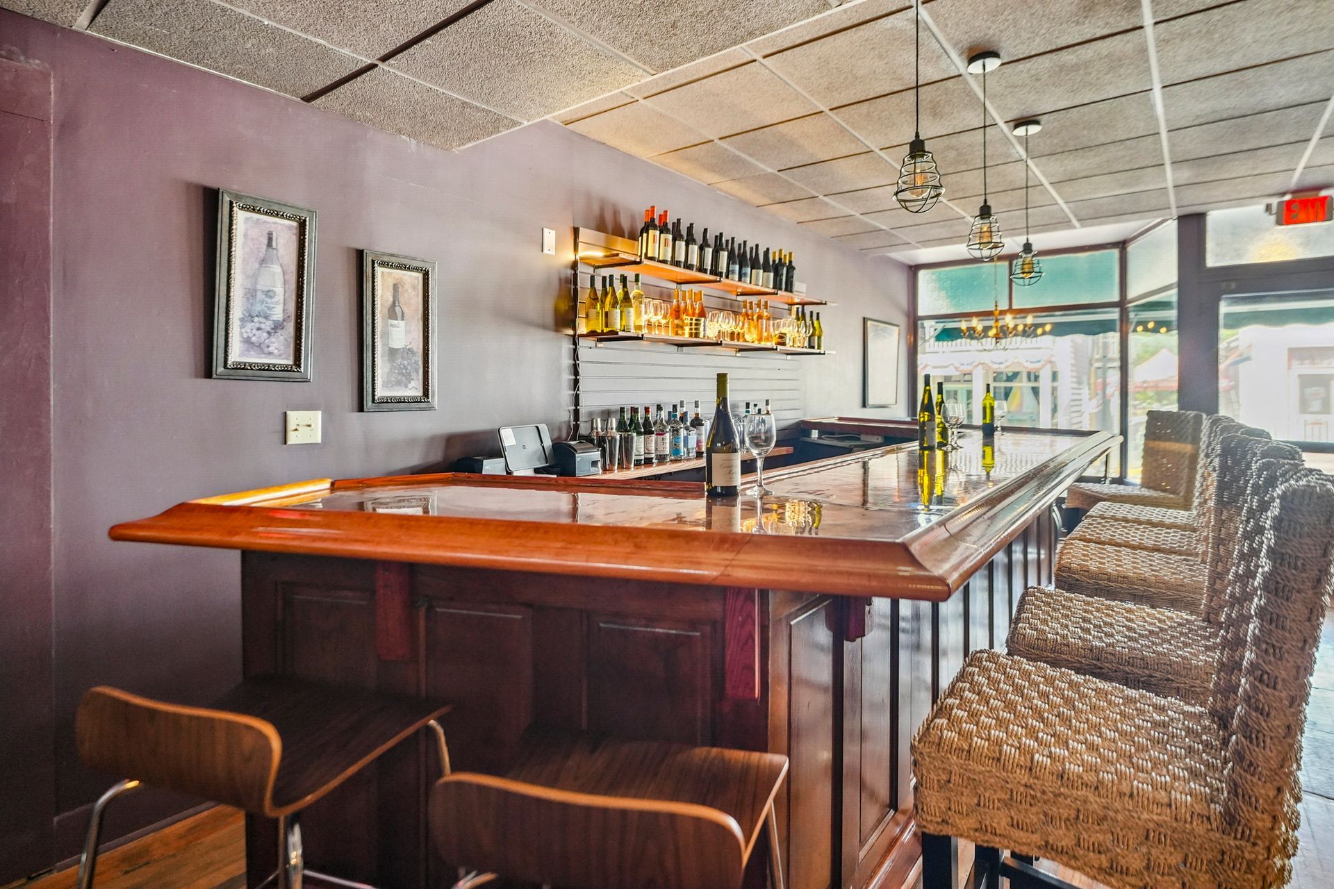 Modern bar interior with wooden counter, woven stools, and bottles on shelves by large windows