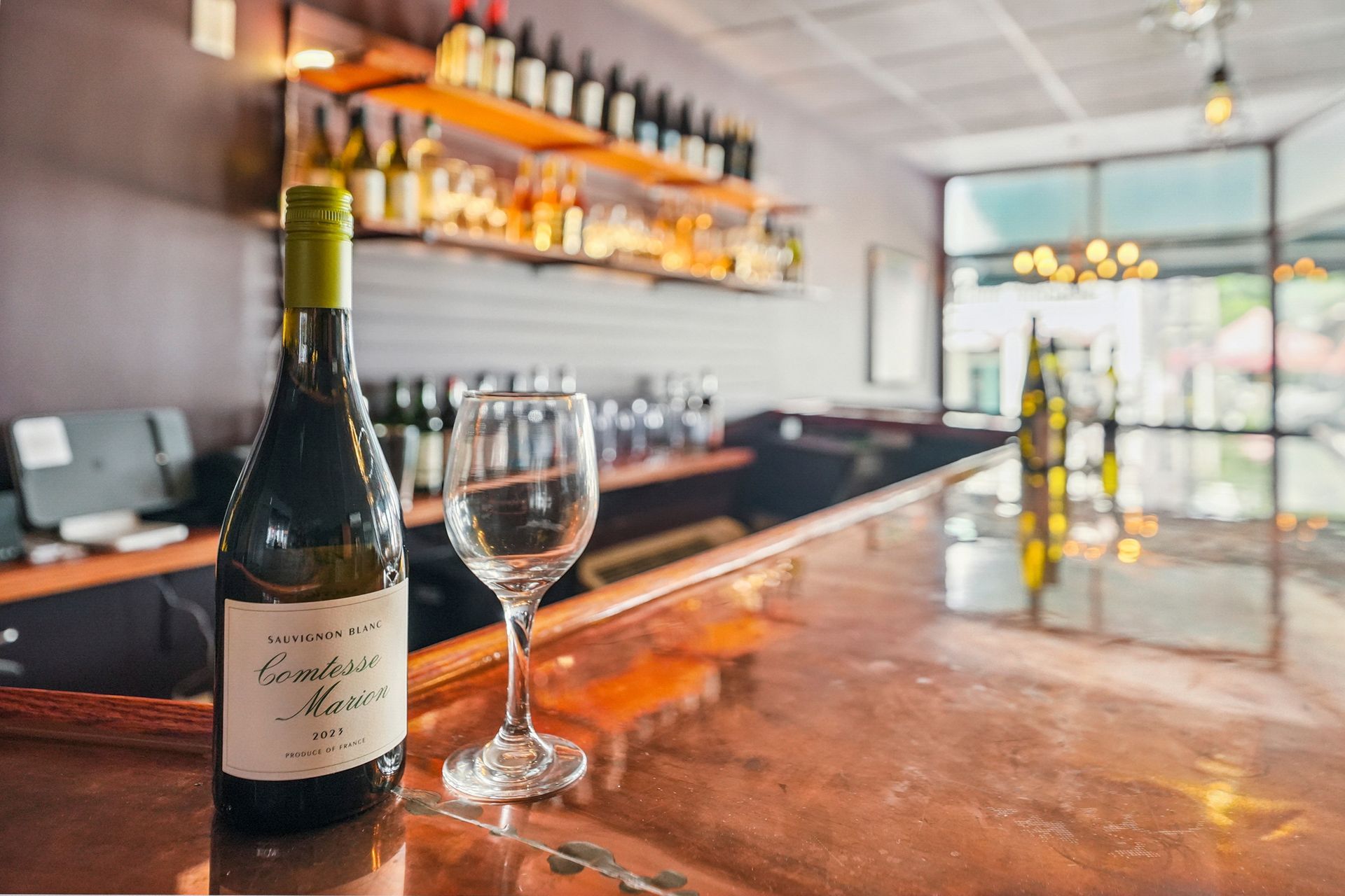 Wine bottle and glass on a bar counter with warm lighting and shelves in the background