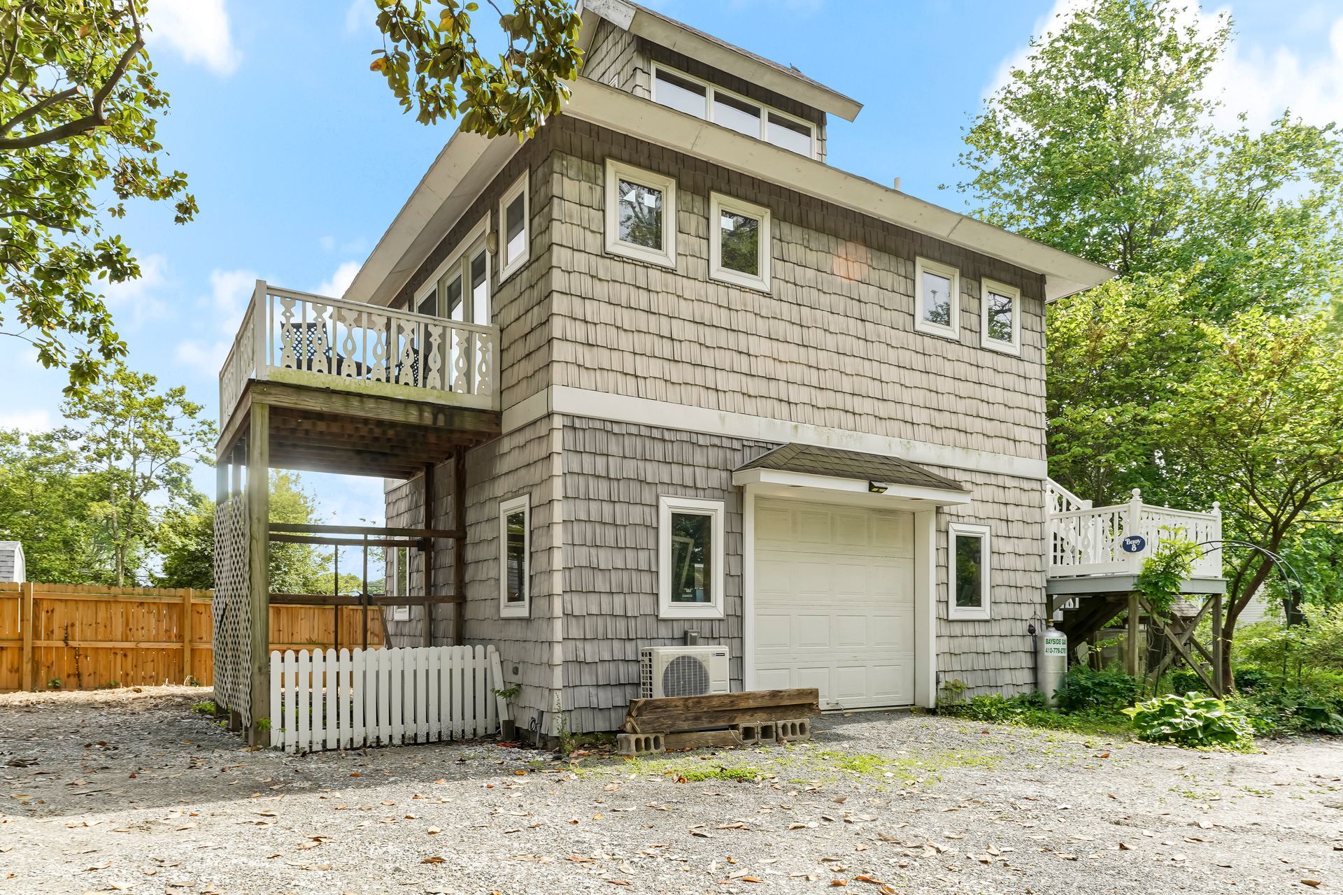 Two-story gray house with garage, balcony, and gravel driveway surrounded by trees