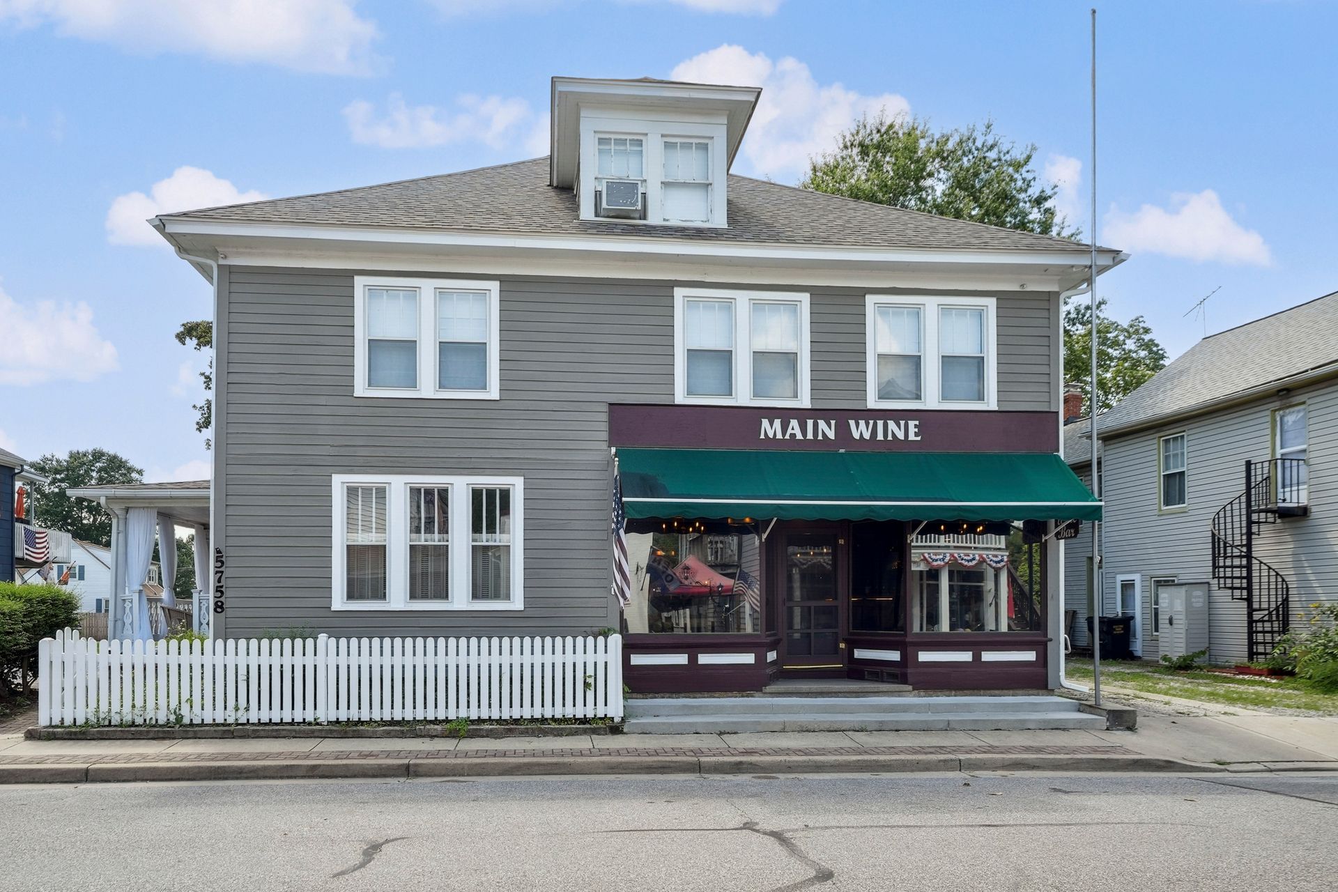 Gray storefront labeled “MAIN WINE” with green awning and white picket fence