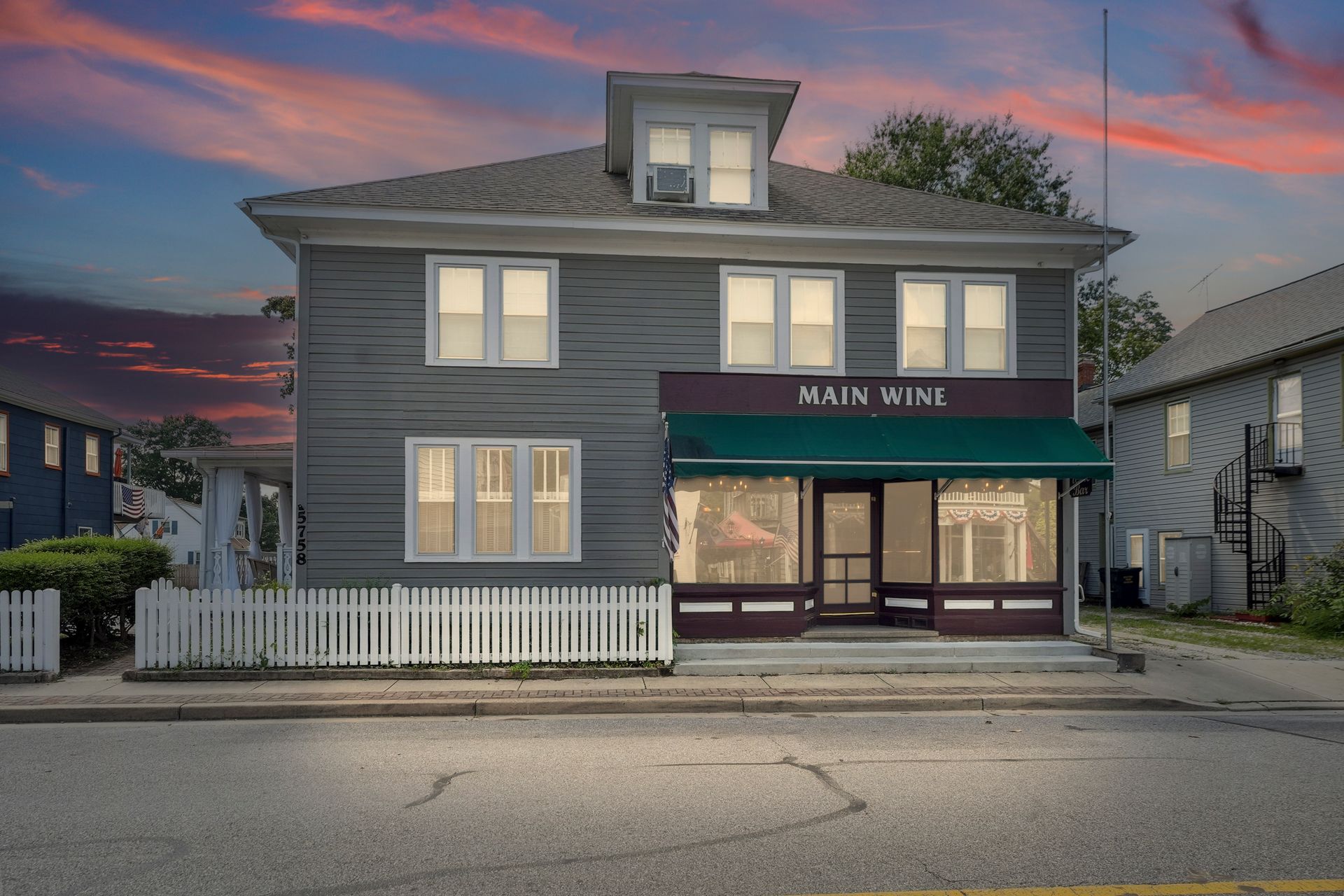 Gray storefront at dusk with white trim, lit windows, and a white picket fence under a pink-blue sky