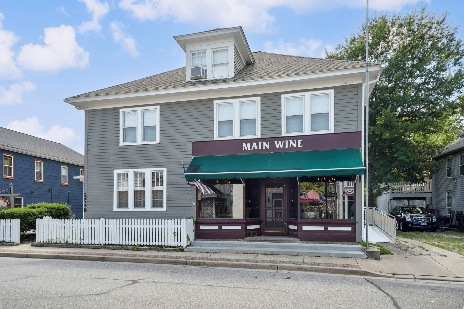 Front view of Main Wine storefront with gray siding, green awning, and white picket fence on a quiet street