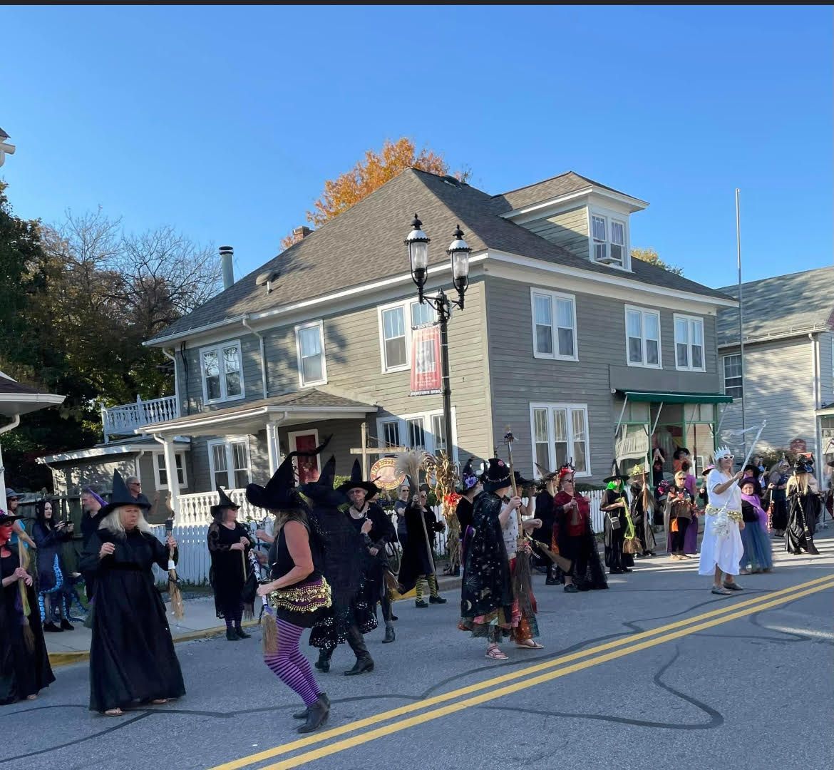 Costumed crowd marches down a street past a gray building on a sunny day