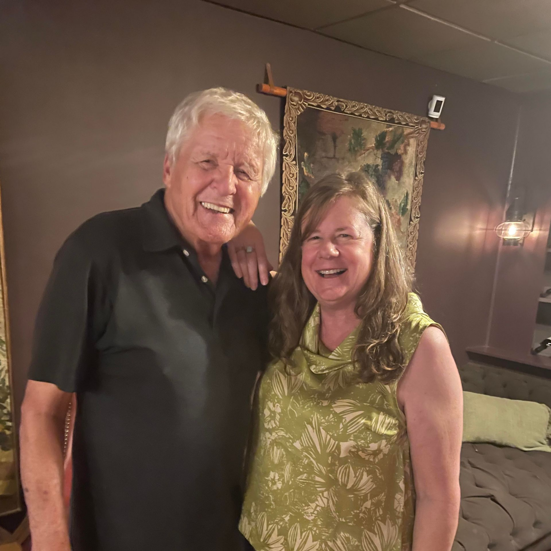 Two smiling people standing together in a warmly lit room, with a bed and wall hanging behind them