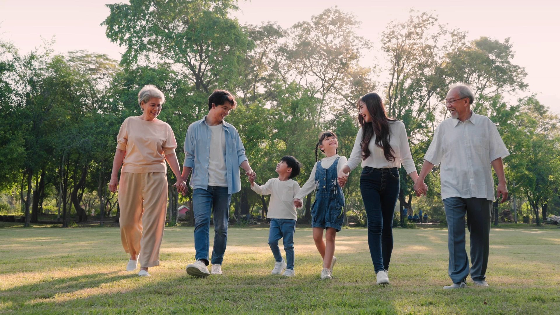 Family of three generations walking in a park, holding hands. Sunlight shines.