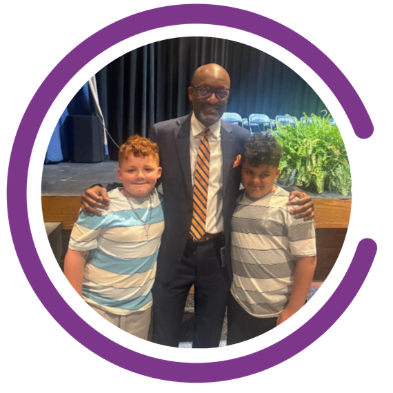Kindness Grows here students and teacher with arms around them. Purple semicircle graphic around the outside of the image