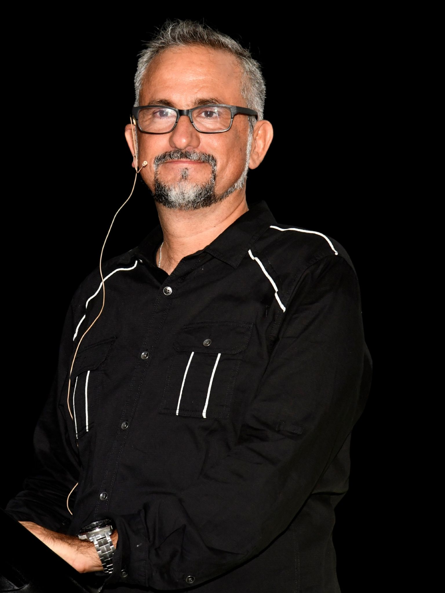 Hombre con camisa negra y gafas, posando frente a un fondo negro y con auriculares puestos.