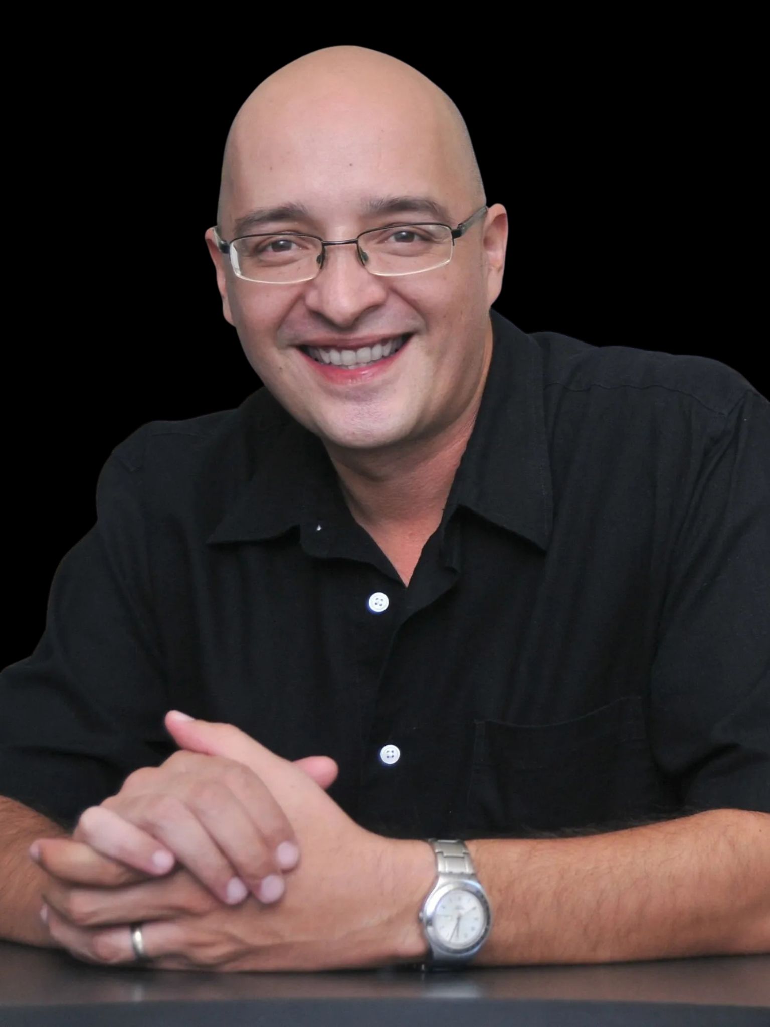 Hombre calvo y sonriente con gafas, camisa negra y las manos entrelazadas, sentado sobre un fondo negro