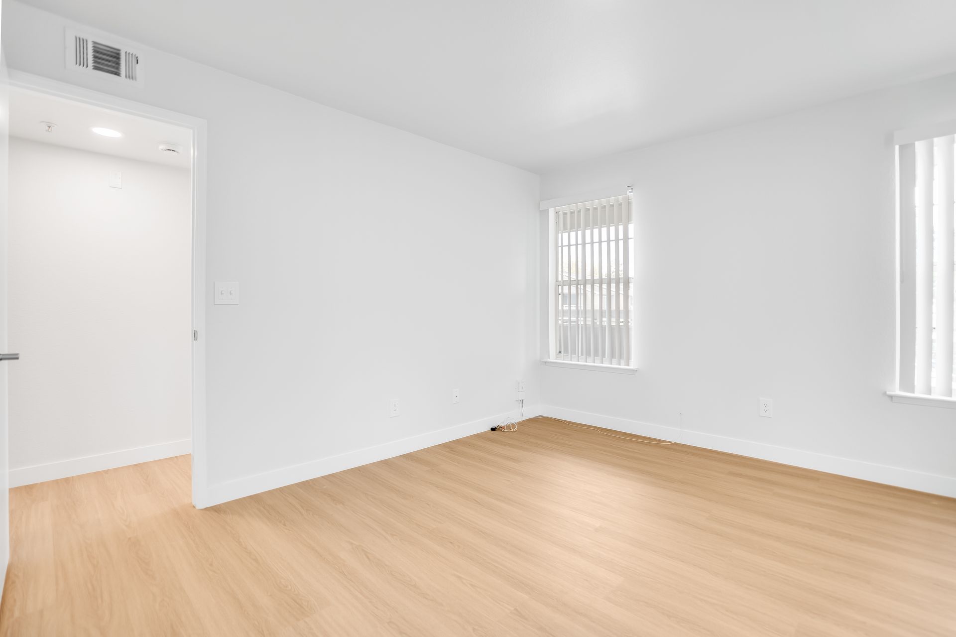 Empty room with light wood floors, white walls, two windows with blinds, and doorway.