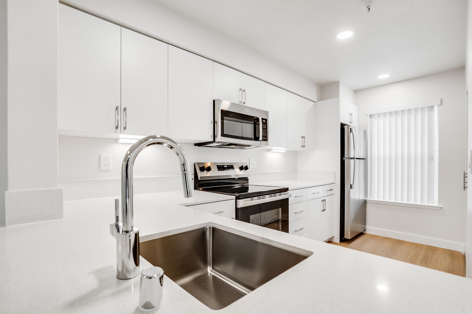 Modern white kitchen with stainless steel appliances, sink, and window with blinds.