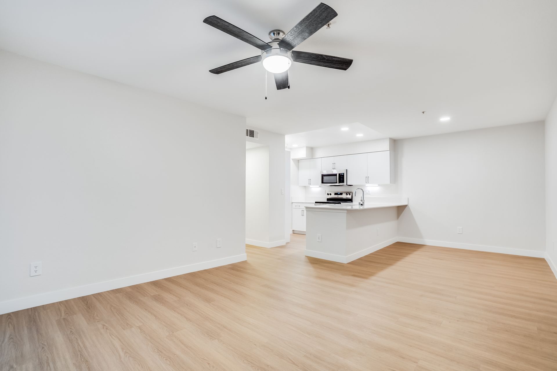 Empty, modern living space with light wood floors, white walls, and a ceiling fan; kitchen visible in the background.