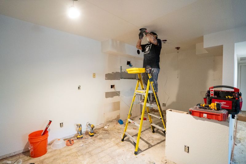 Man on a ladder installing a light fixture in a room under construction, tools and materials present.