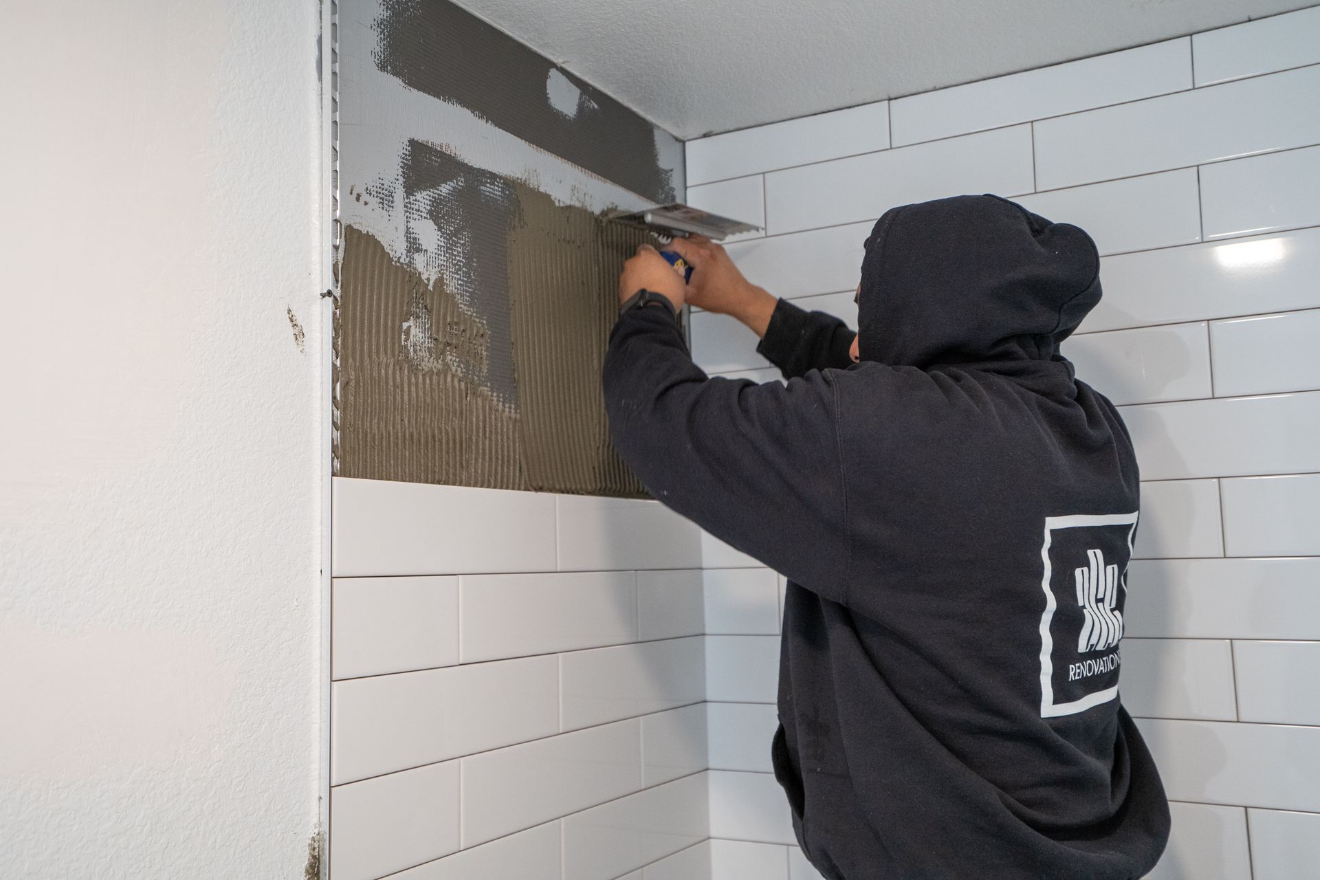 Person tiling a white subway tile wall in a bathroom, applying mortar with a trowel.