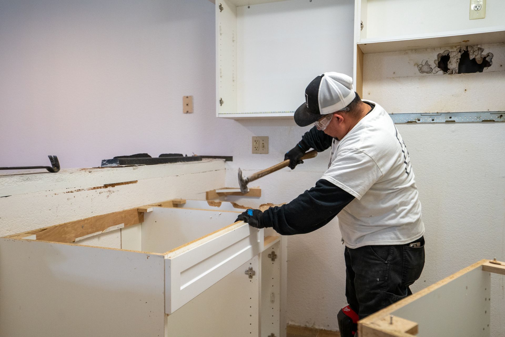 Person in a kitchen removing cabinets with a hammer, wearing gloves and safety glasses.