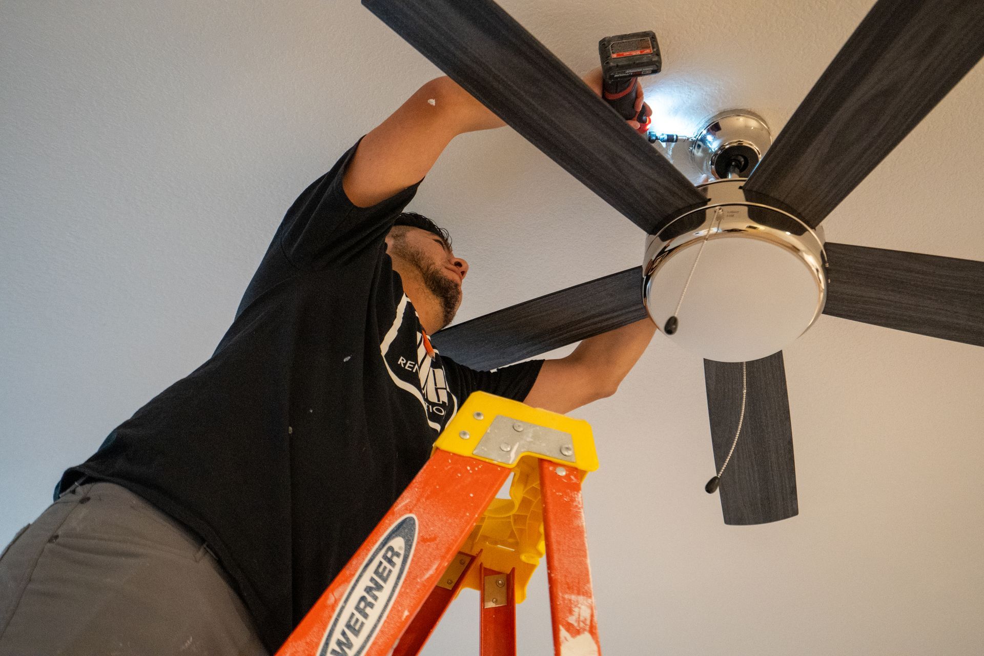 Person installing a ceiling fan, using a power drill while standing on a step ladder.